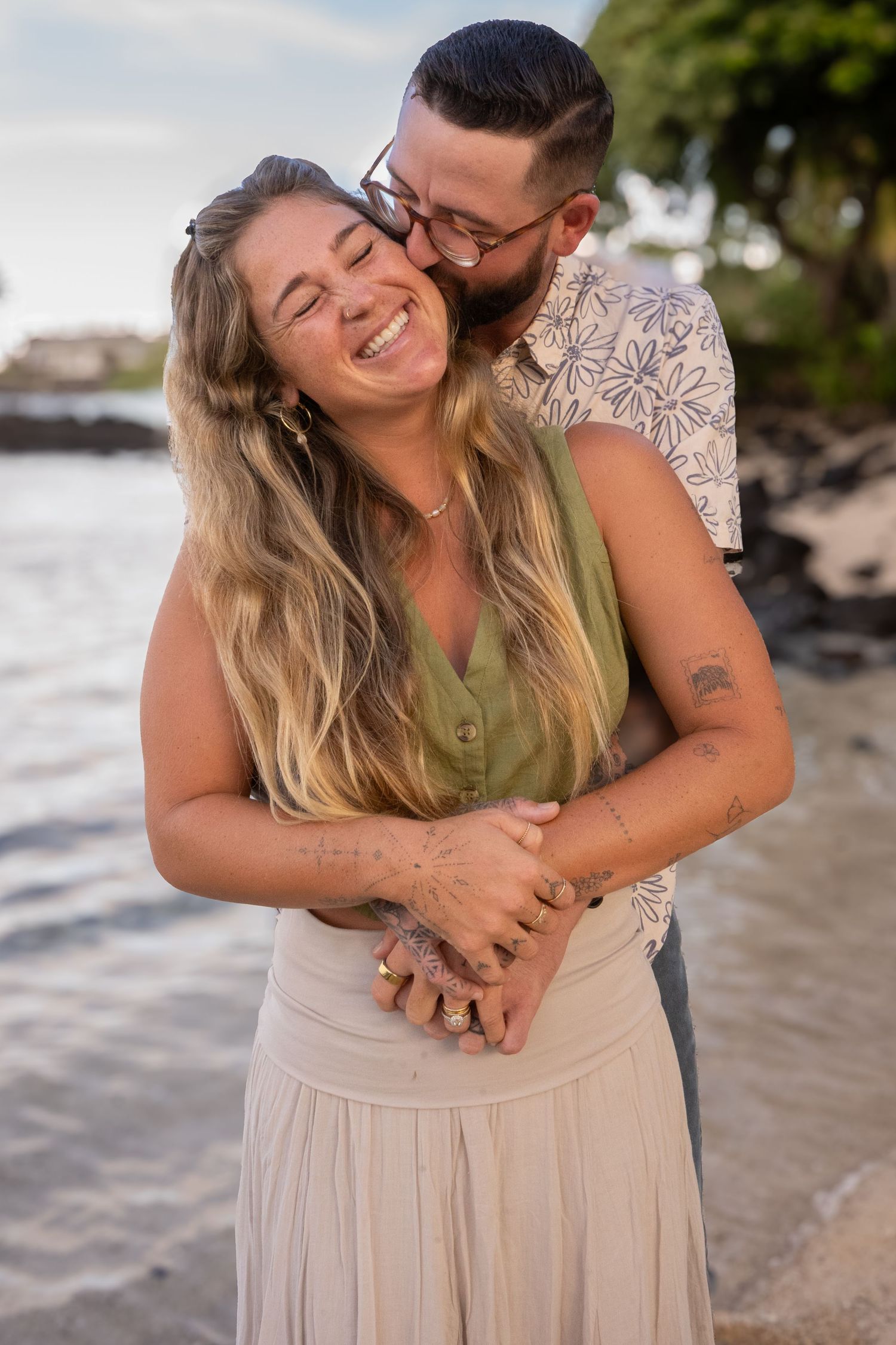 A couple shares an intimate embrace on a beach at sunset while wearing casual beige and olive green outfits.