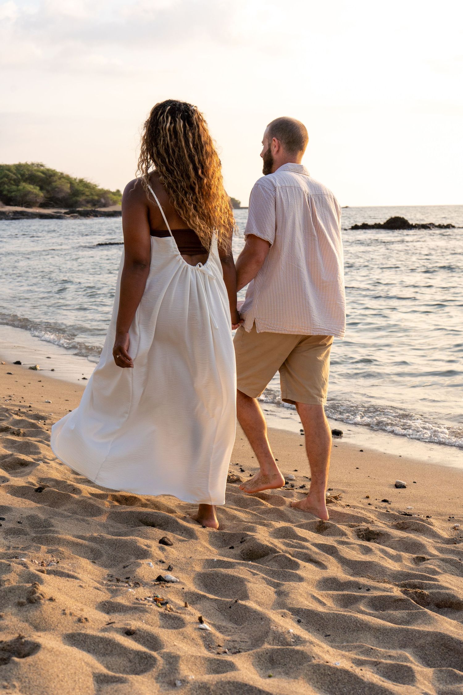 Couple walking hand in hand on a sandy beach at sunset, woman in white dress, man in casual shorts and shirt.