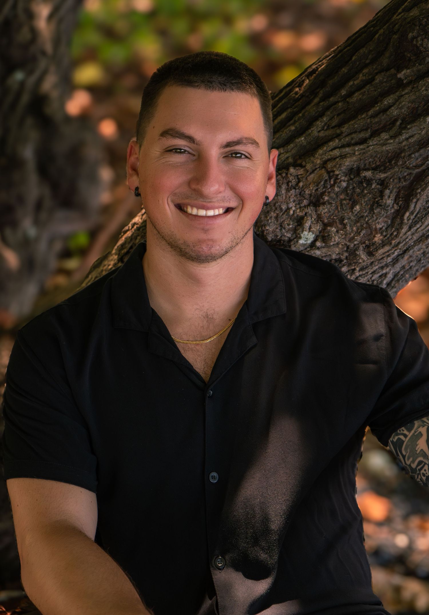 Man in black shirt smiling while leaning against tree trunk.