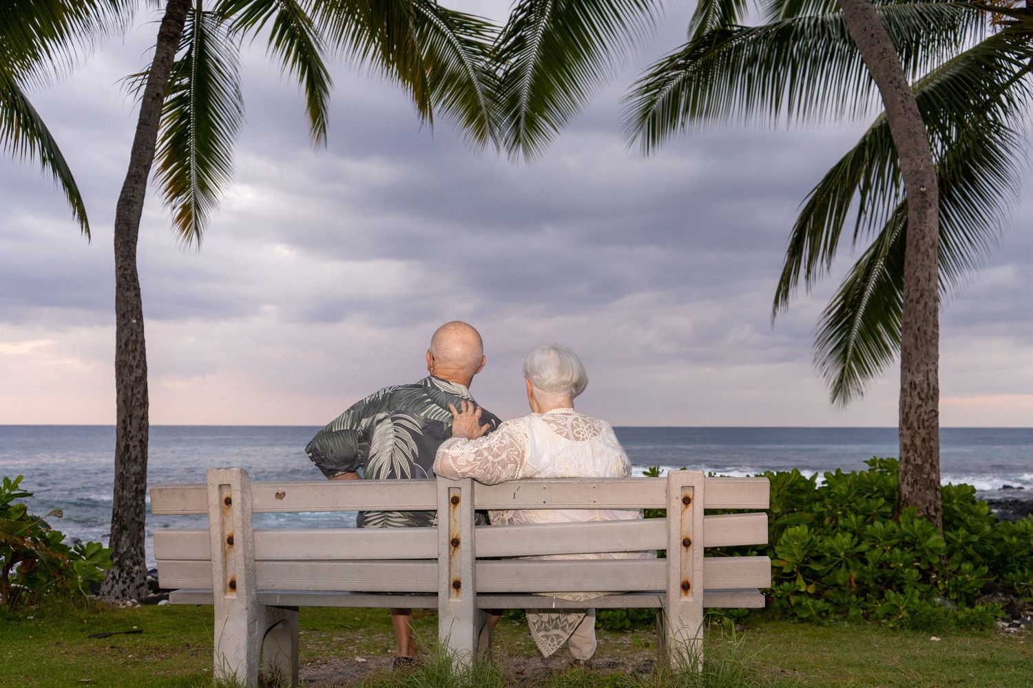 Elderly couple sitting on a wooden bench between palm trees, gazing at the ocean under a cloudy sky.