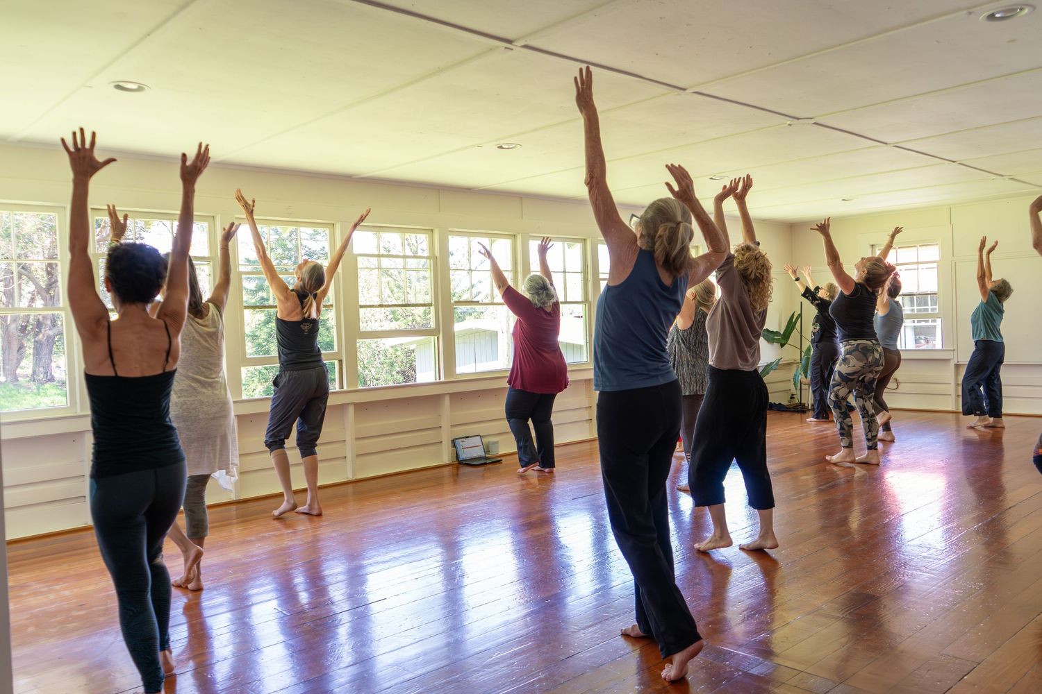 A group of people stretching with raised arms during a fitness class in a bright studio with wooden floors and mirrors.