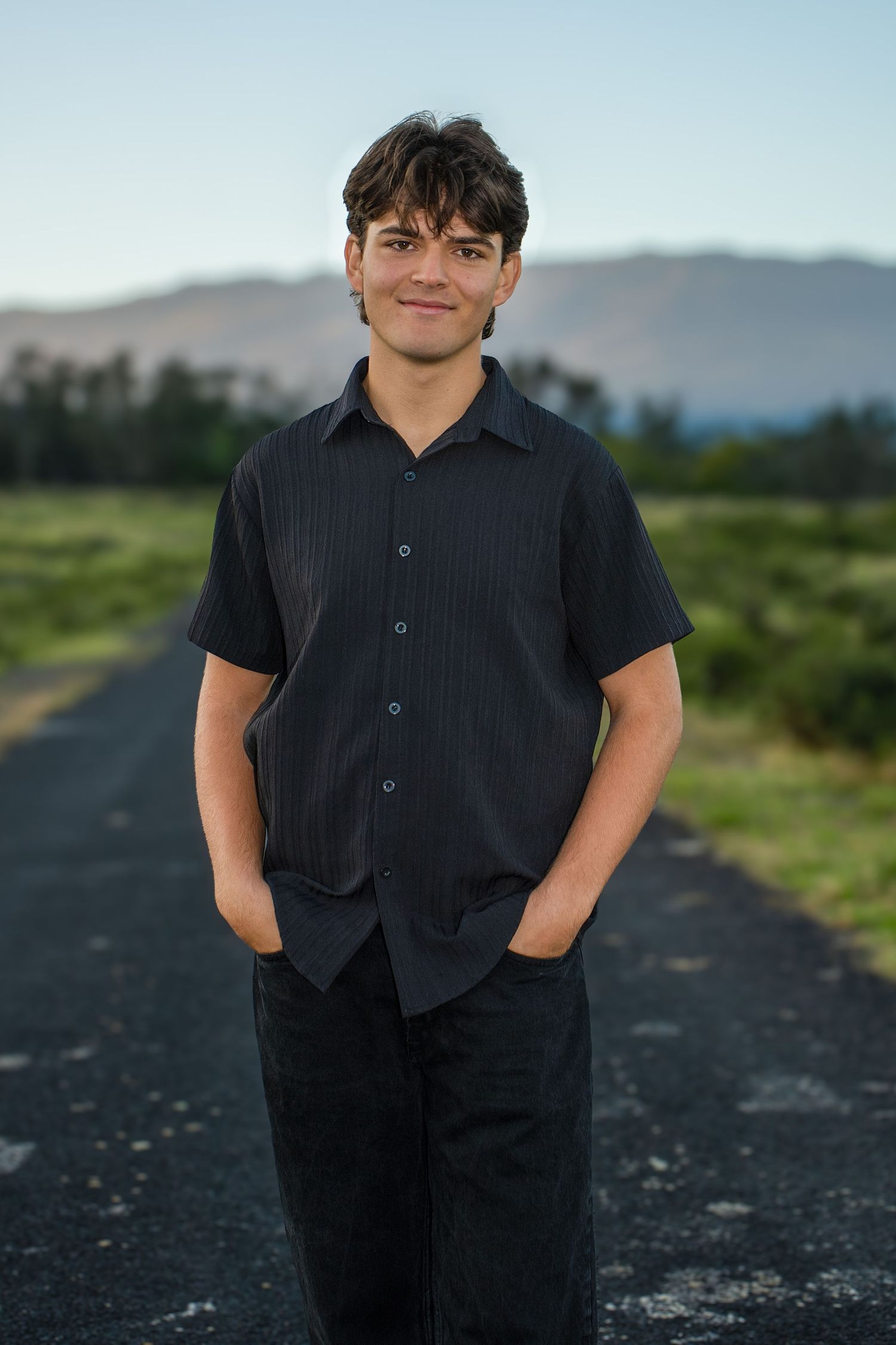 A figure in a dark shirt stands on a gravel path with mountains and greenery visible in the background.