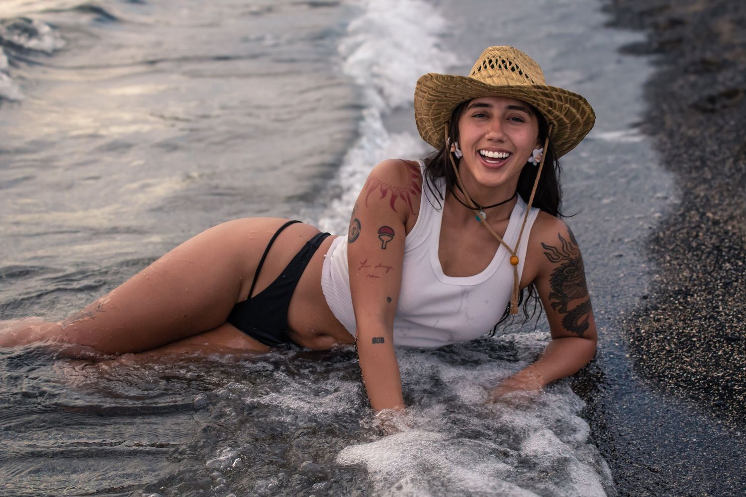 Person in beachwear and straw hat plays in ocean waves on black sand beach.