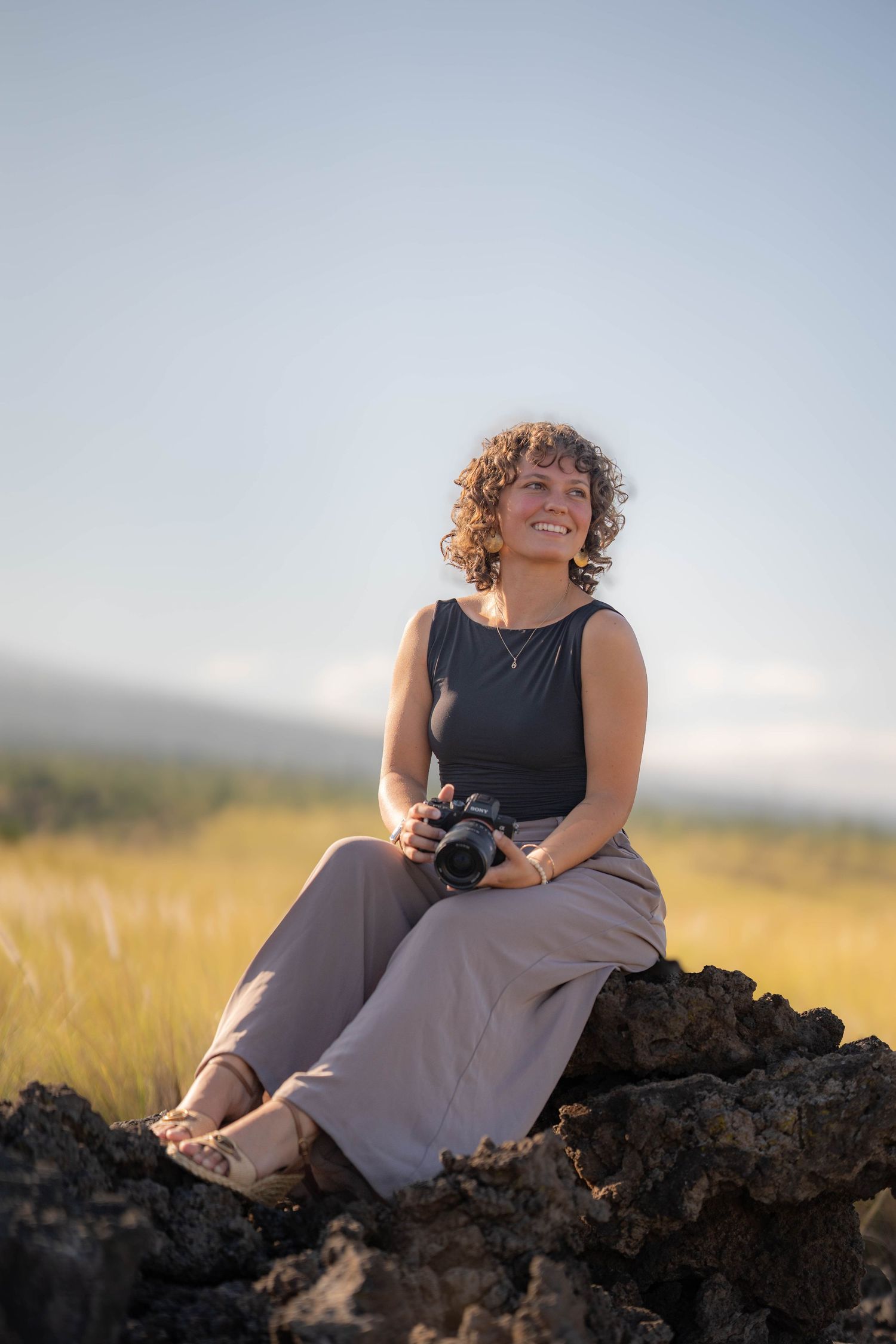 A smiling woman with curly hair sits on rocks in a field, holding a camera, wearing a black top and gray pants.