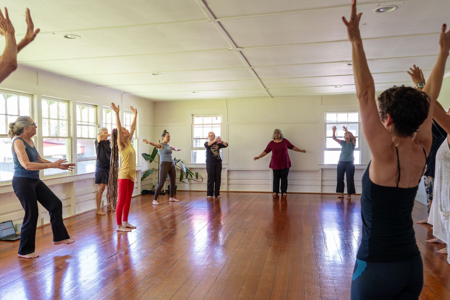 Group fitness class participants stretch with arms raised in a bright studio with wooden floors and natural light.