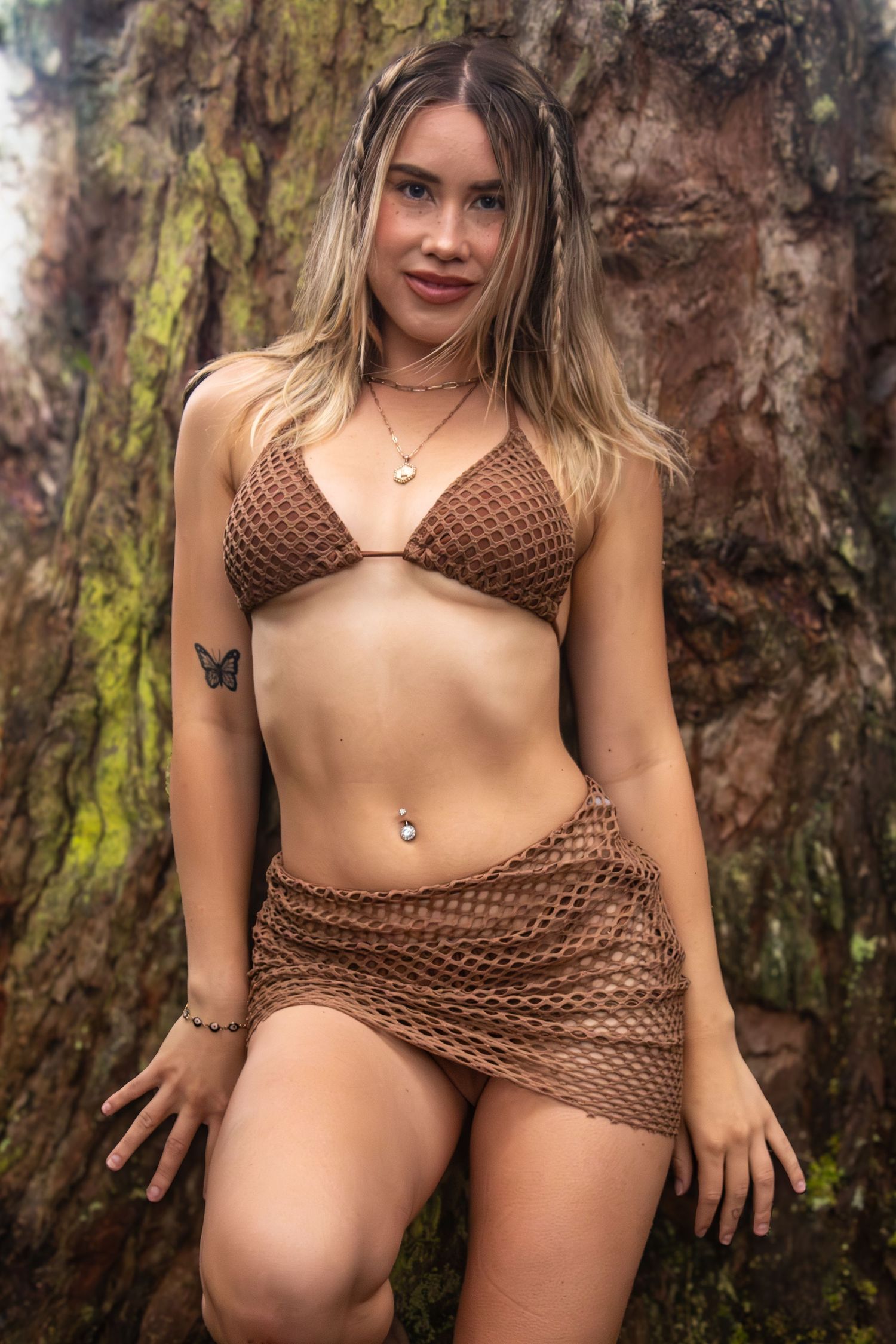 Model in brown crochet beachwear poses against mossy tree bark in natural setting.
