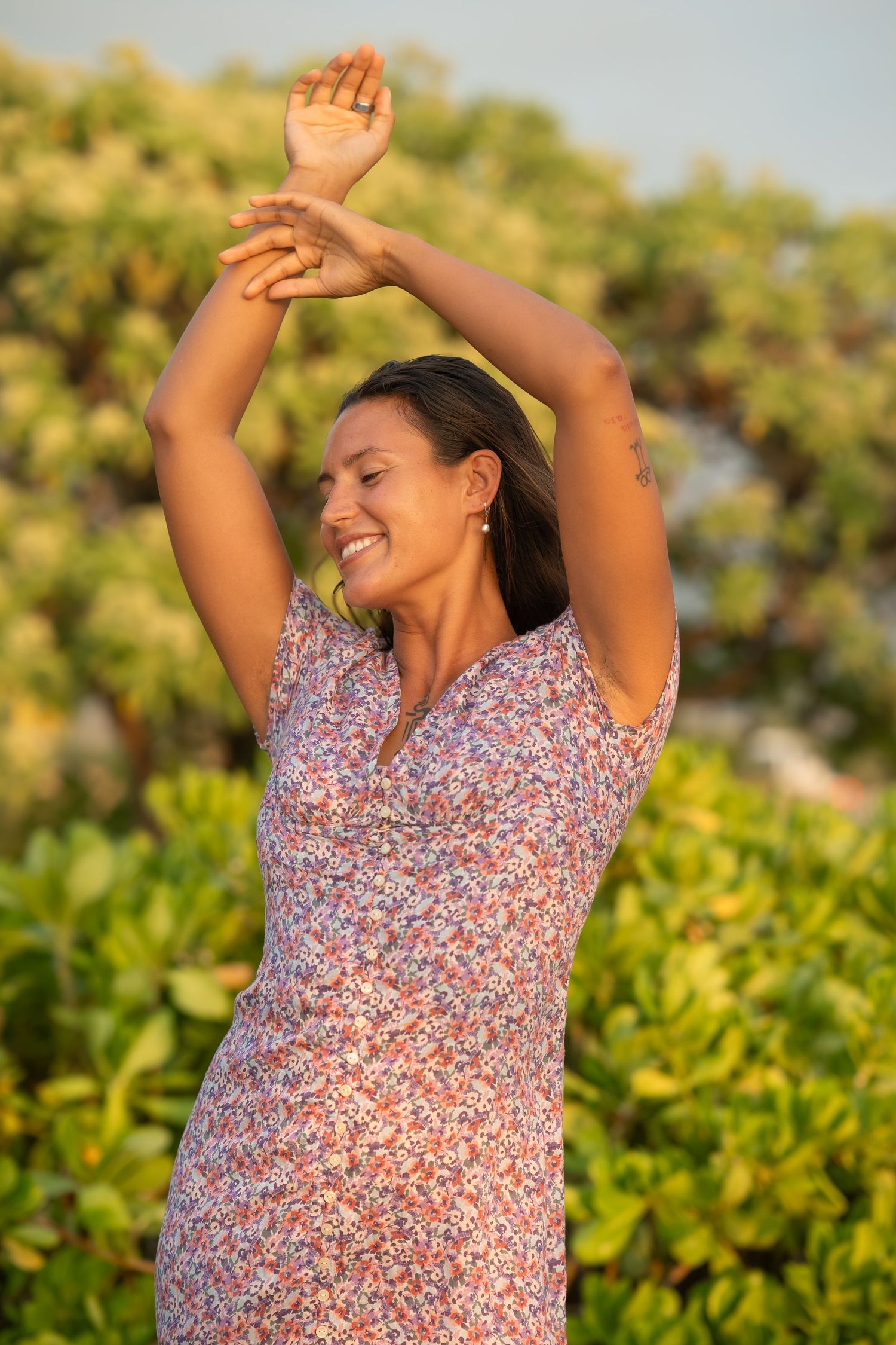 Woman in floral dress dances joyfully outdoors against green foliage in warm sunlight.