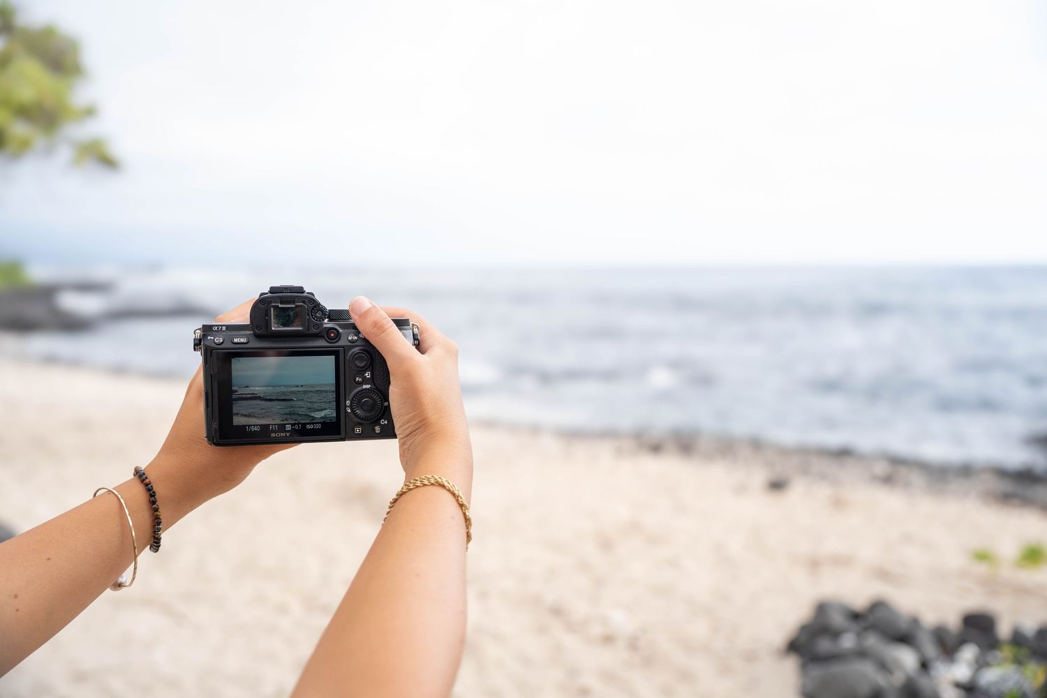 Person holding a DSLR camera on a tropical beach, capturing ocean views on a sunny day in Hawaii.