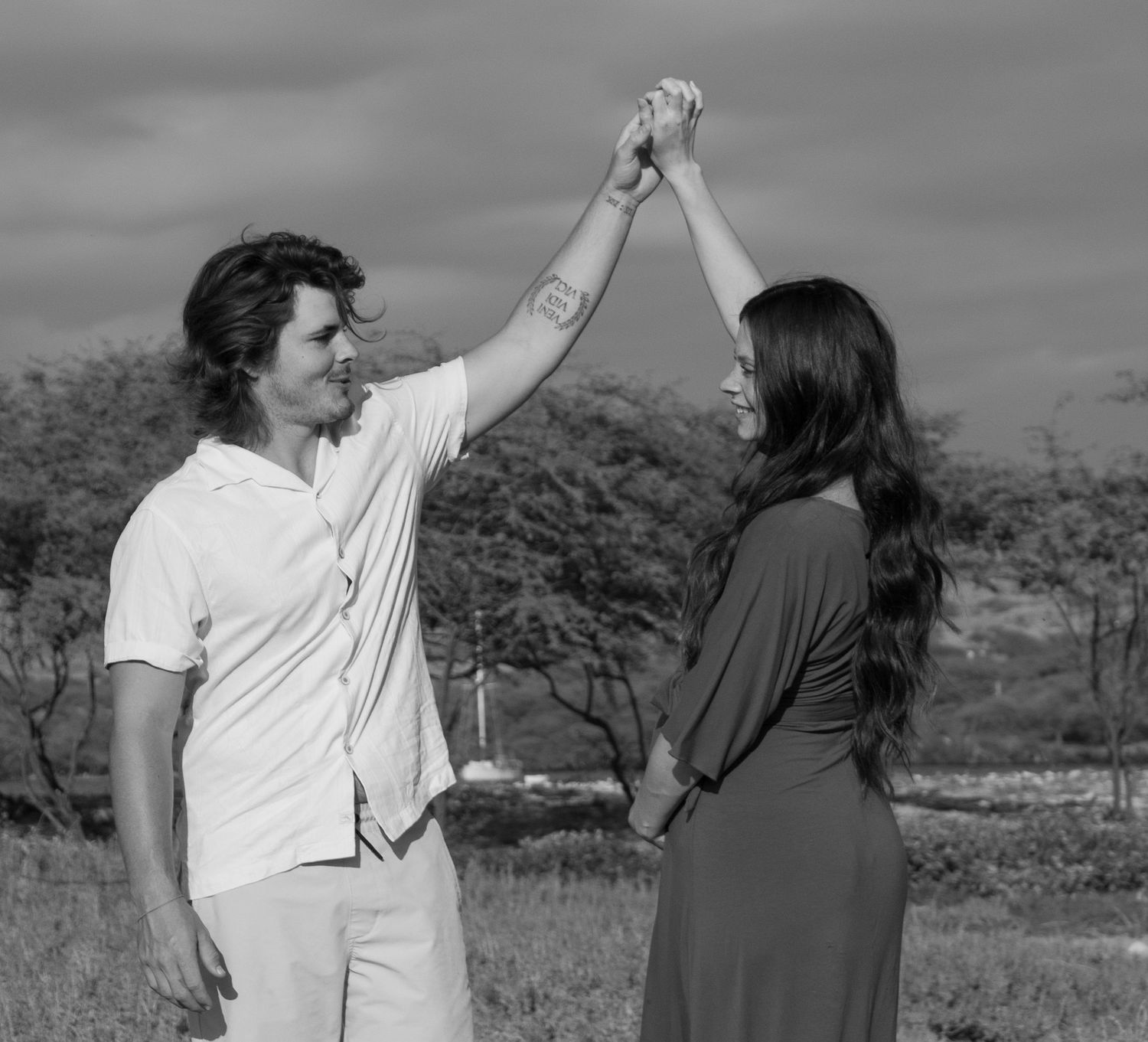 A couple in casual attire dance together outdoors against a natural landscape with trees in black and white.