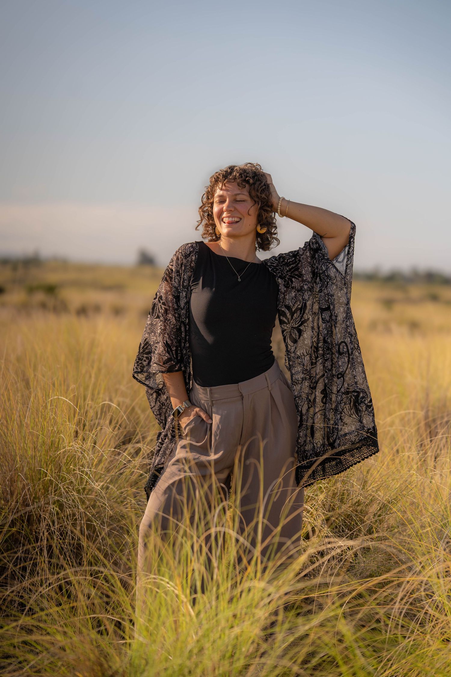 Smiling woman with curly hair in black top and sheer kimono stands in golden grassland at sunset.