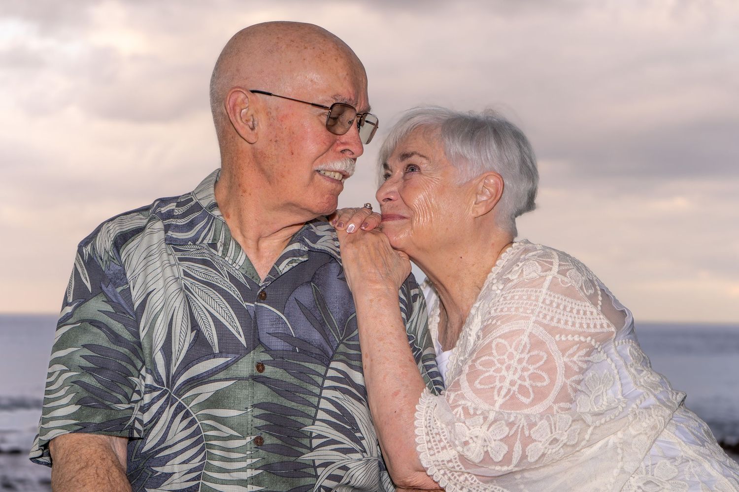 Elderly couple sharing a romantic moment at the beach during sunset, man in Hawaiian shirt kissing woman's hand.