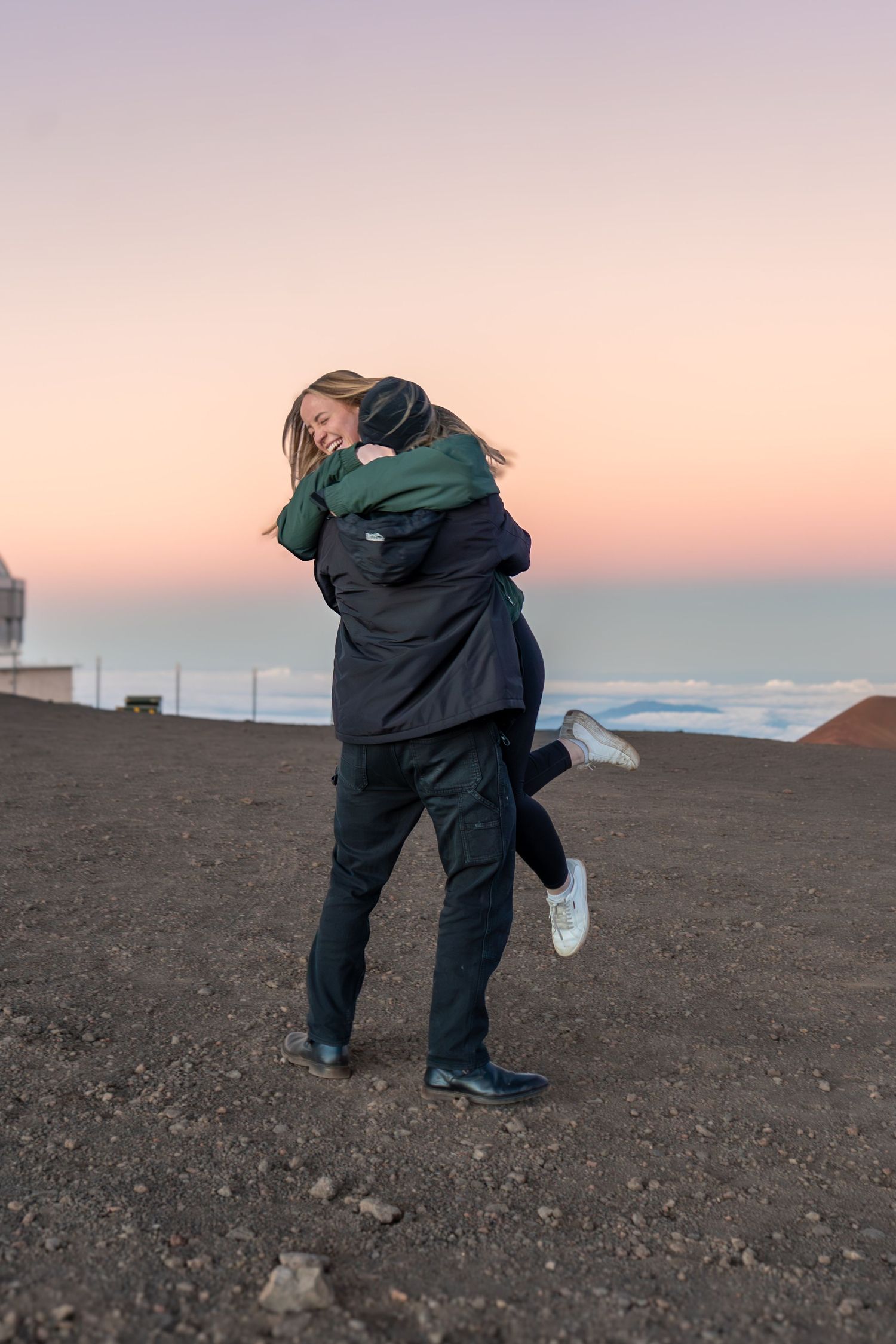 Couple embracing at sunset on a volcanic summit, woman lifted off ground, clouds below horizon.