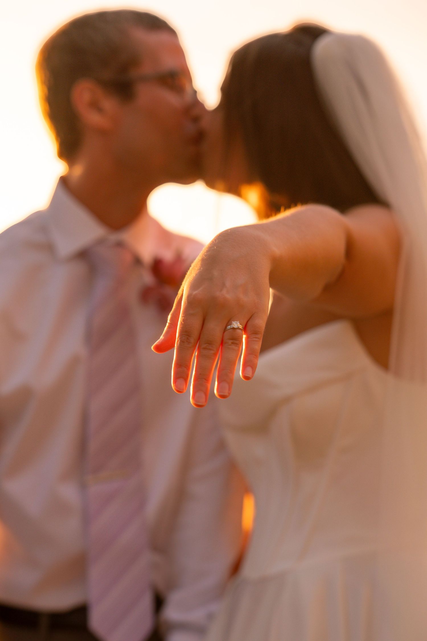 Bride and groom kissing at sunset while bride shows off her diamond wedding ring to the camera.