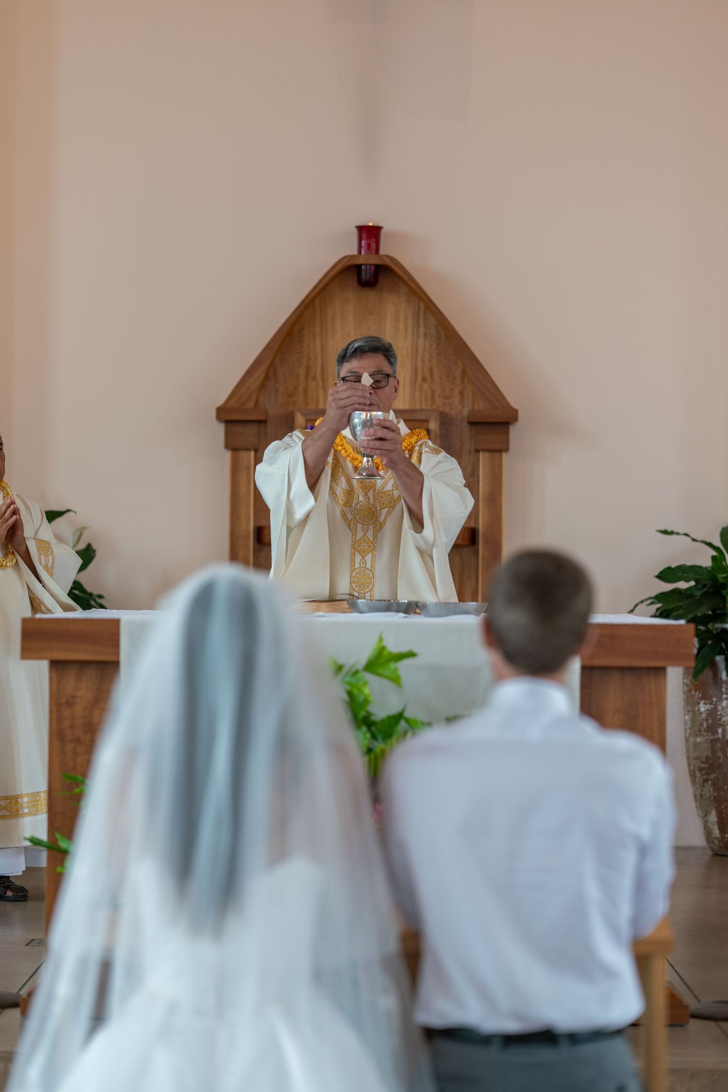 Priest elevating chalice at altar during Catholic wedding ceremony with bride and groom kneeling before him.