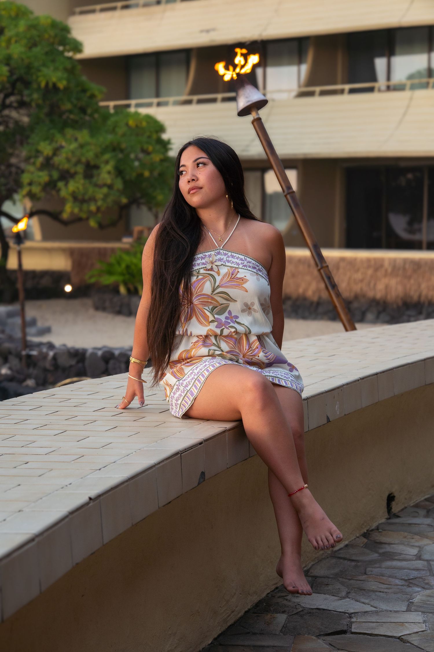 Young woman in floral strapless dress sits barefoot on a curved wall at a Hawaiian resort during golden hour.