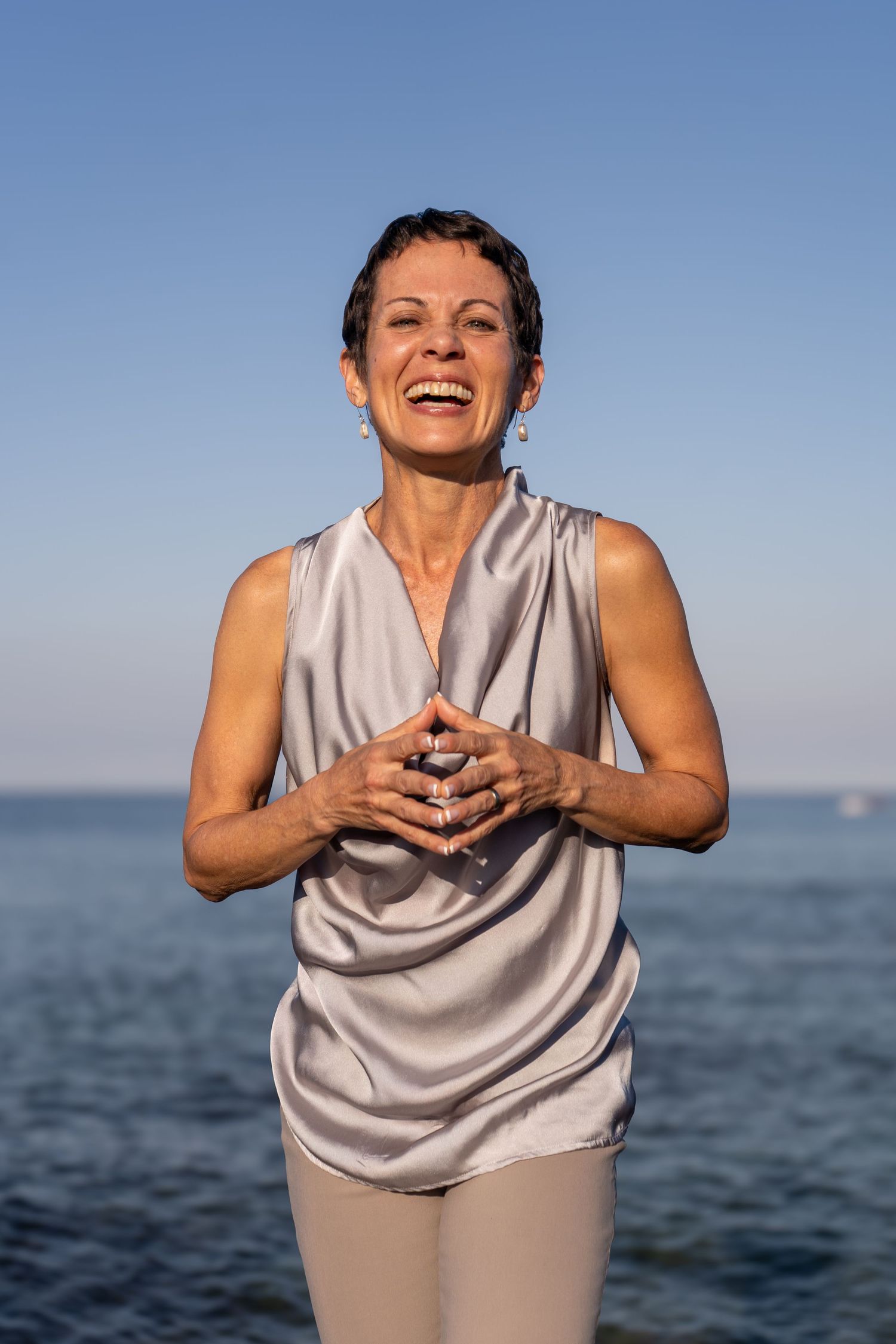 A person in a light gray sleeveless top laughs joyfully against an ocean backdrop in a series of expressive poses.