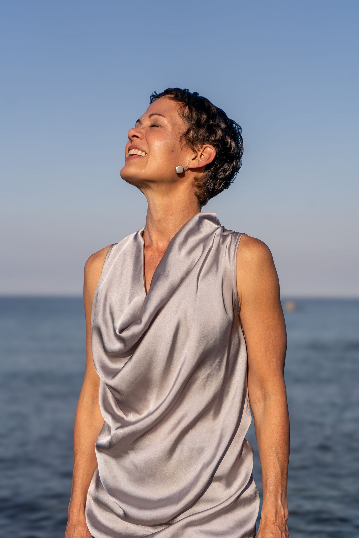 Person in sleeveless gray top laughs joyfully against ocean backdrop during golden hour.
