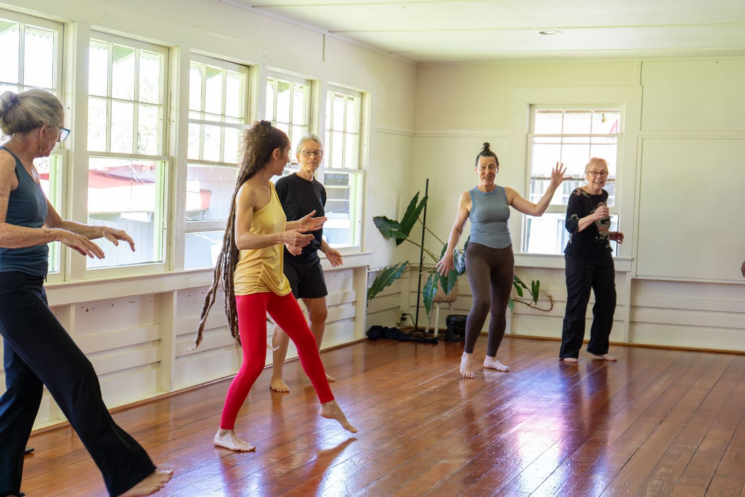 Group fitness class participants exercise together in a bright studio with hardwood floors and large windows.