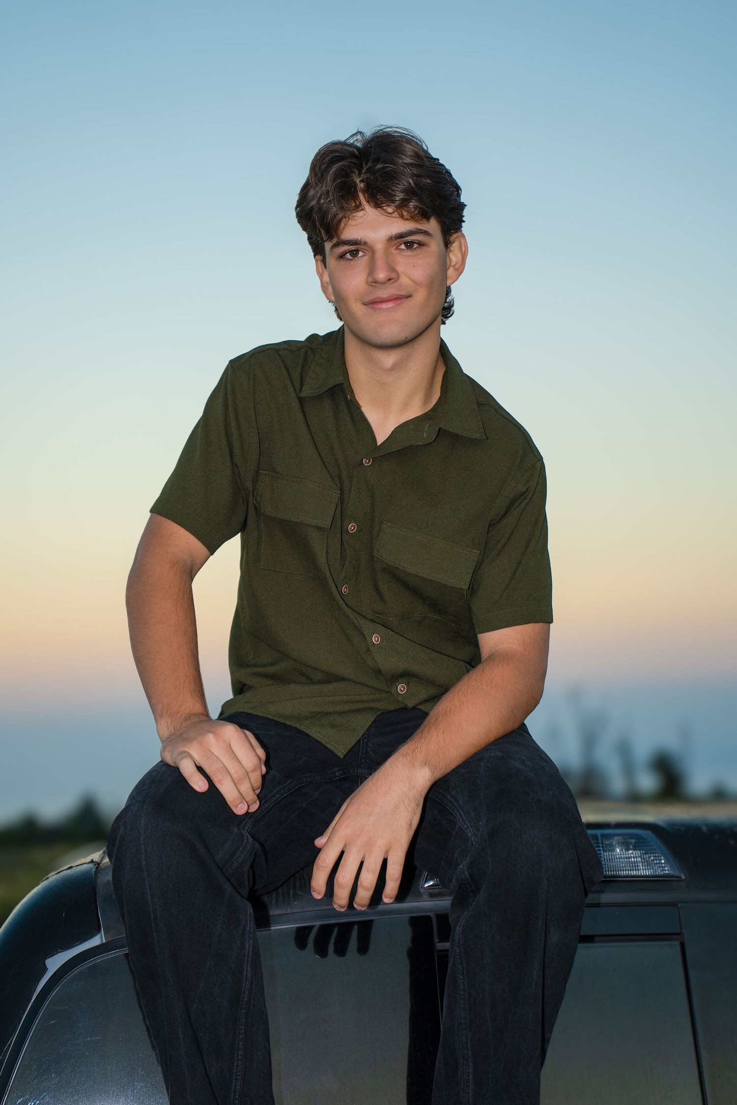 Young man in green shirt sitting on car hood at dusk, smiling confidently outdoors.