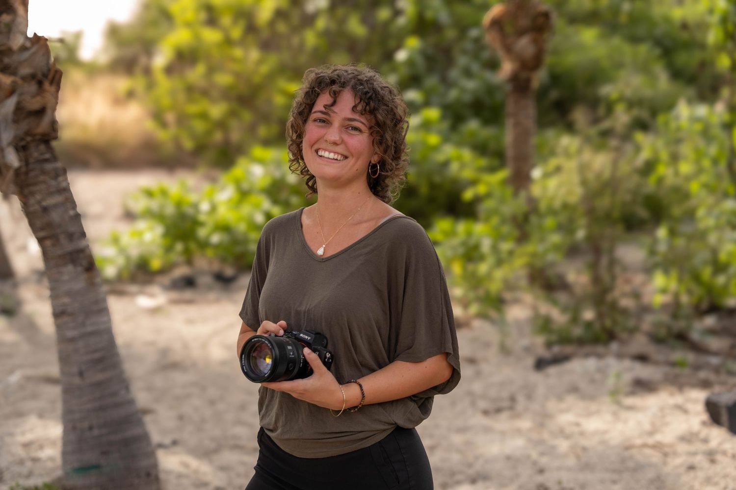 Smiling woman with curly hair holding a DSLR camera outdoors in a tropical setting with palm trees.