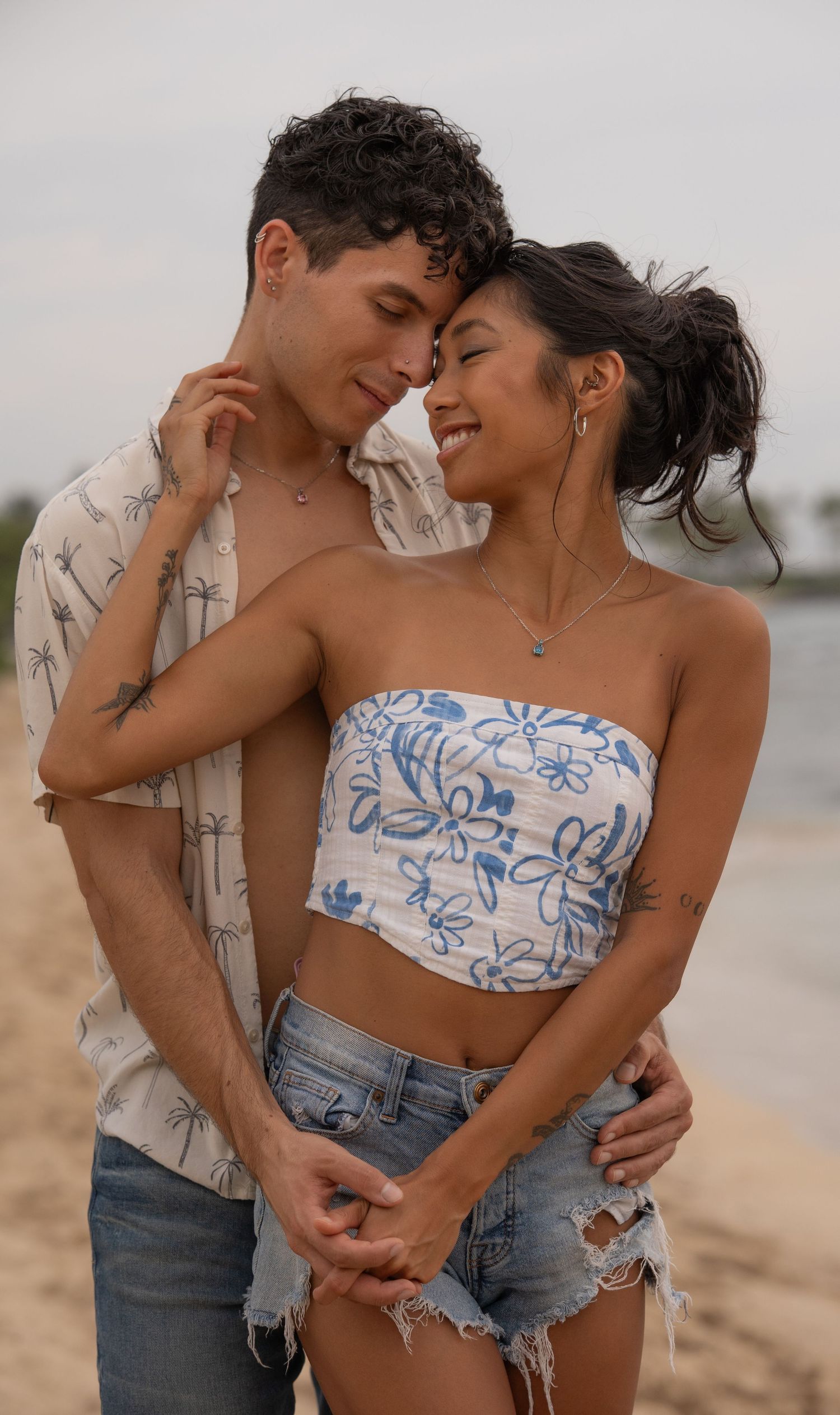 A couple embraces on a sandy beach, woman in floral tube top and denim shorts, man in palm tree print shirt.