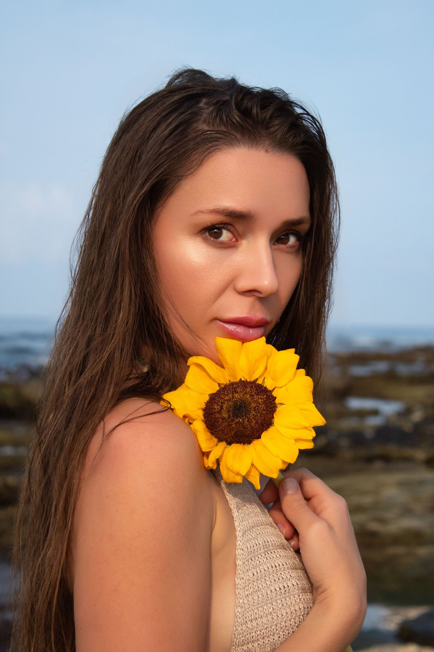 Close-up portrait with bright yellow sunflower against blue sky and ocean backdrop.