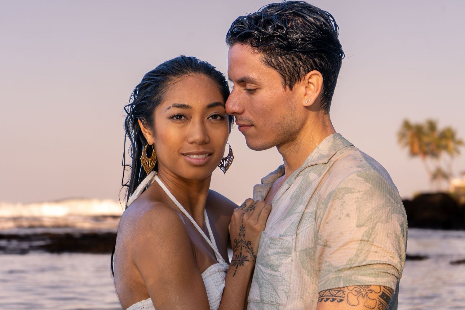 Romantic couple embraces at sunset on a tropical beach with palm trees and ocean views in the background.