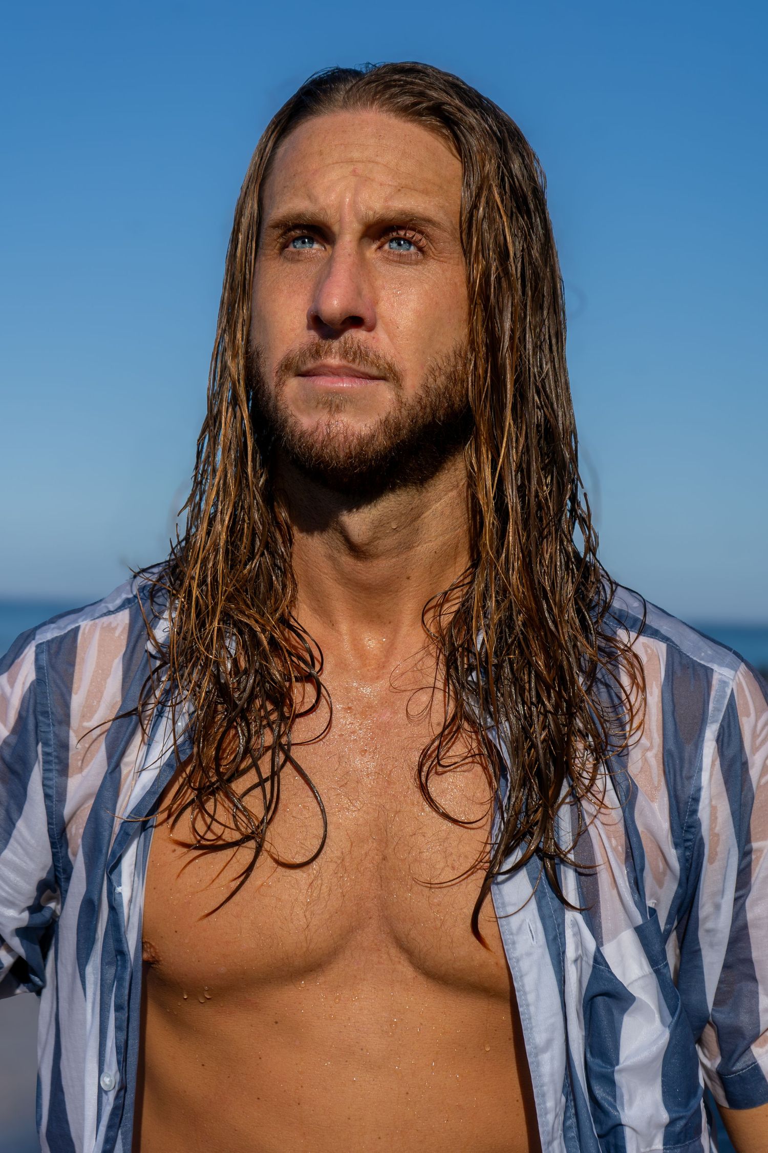 Man with wet long hair wearing striped shirt against blue sky background.