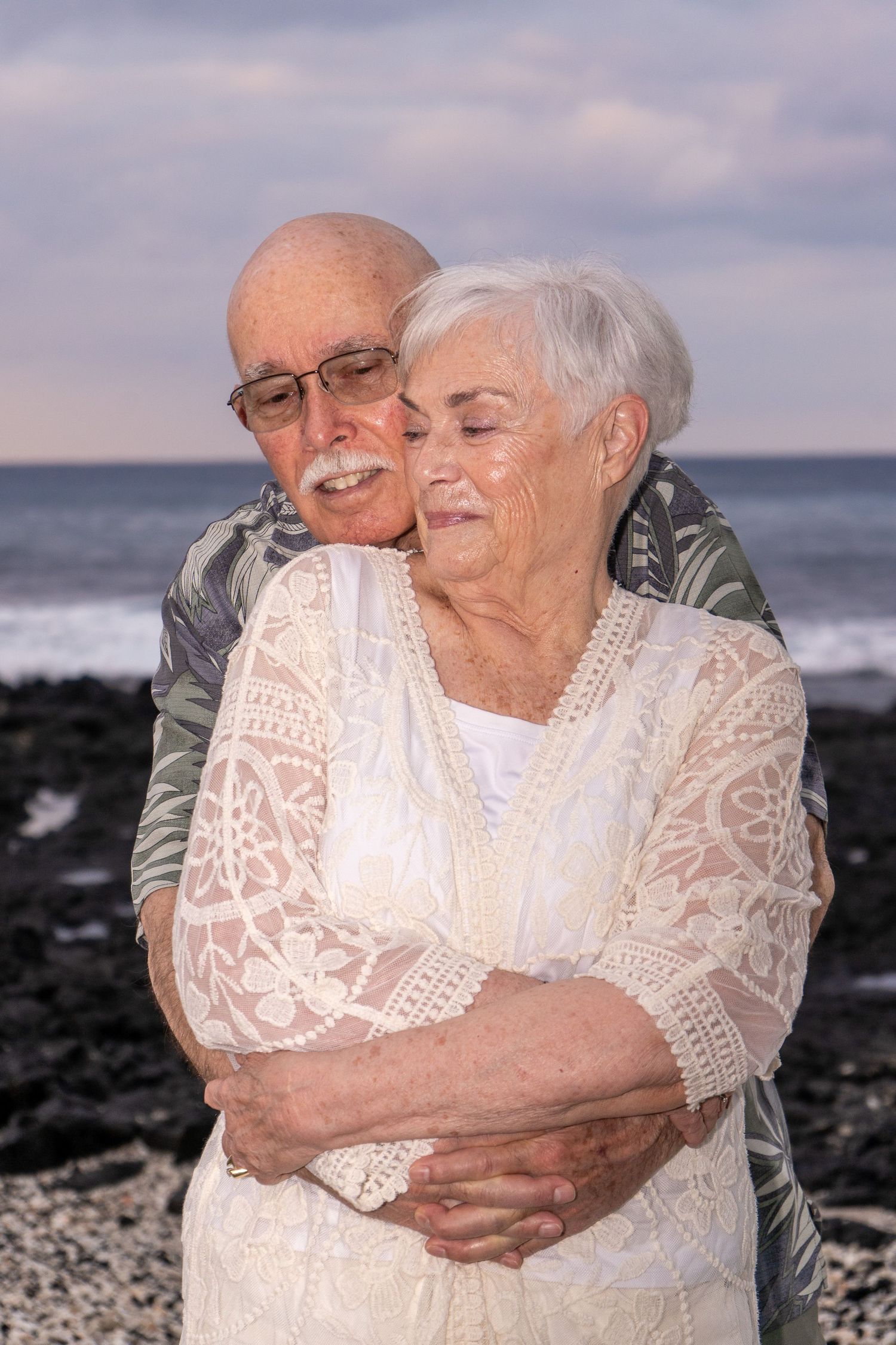 Elderly couple embracing on rocky beach, woman in white lace dress, man in floral shirt, ocean waves behind them.