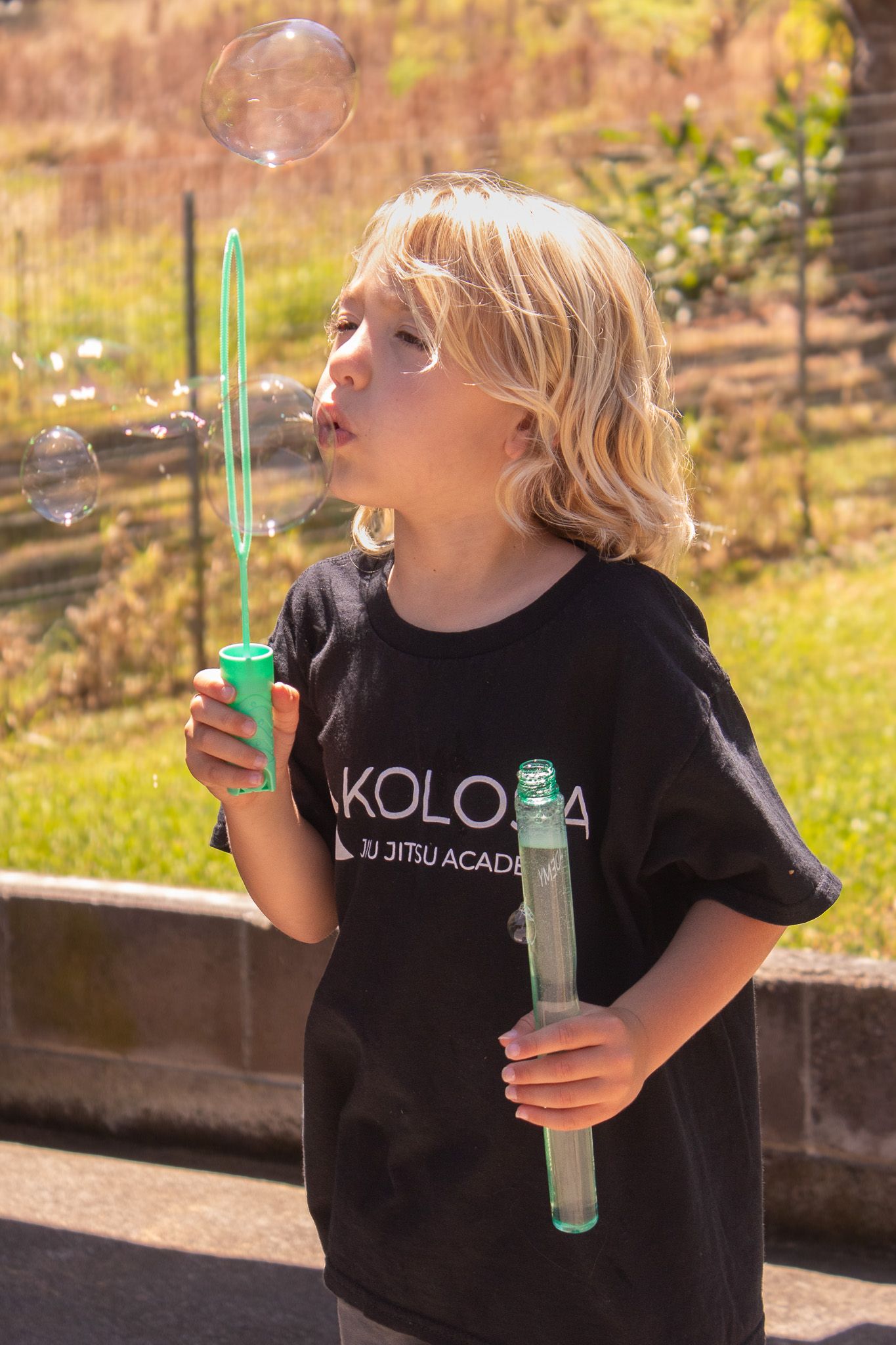 Child in black t-shirt blowing bubbles outdoors on sunny day.