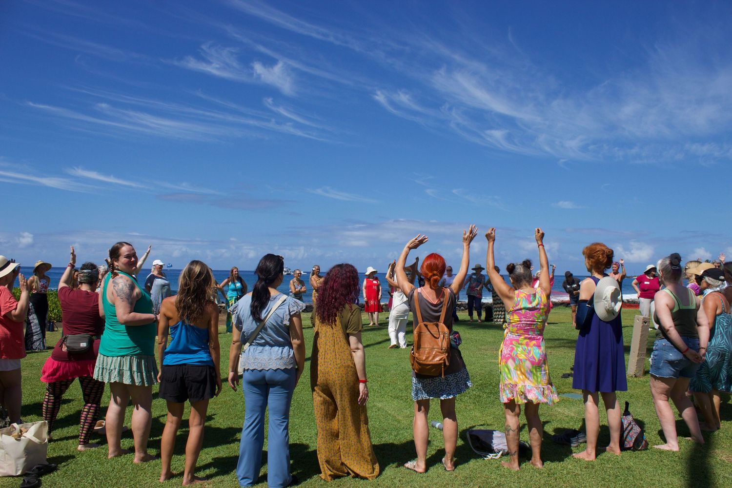 Group of women gathered outdoors on green grass raising hands under a bright blue sky at a festival event.