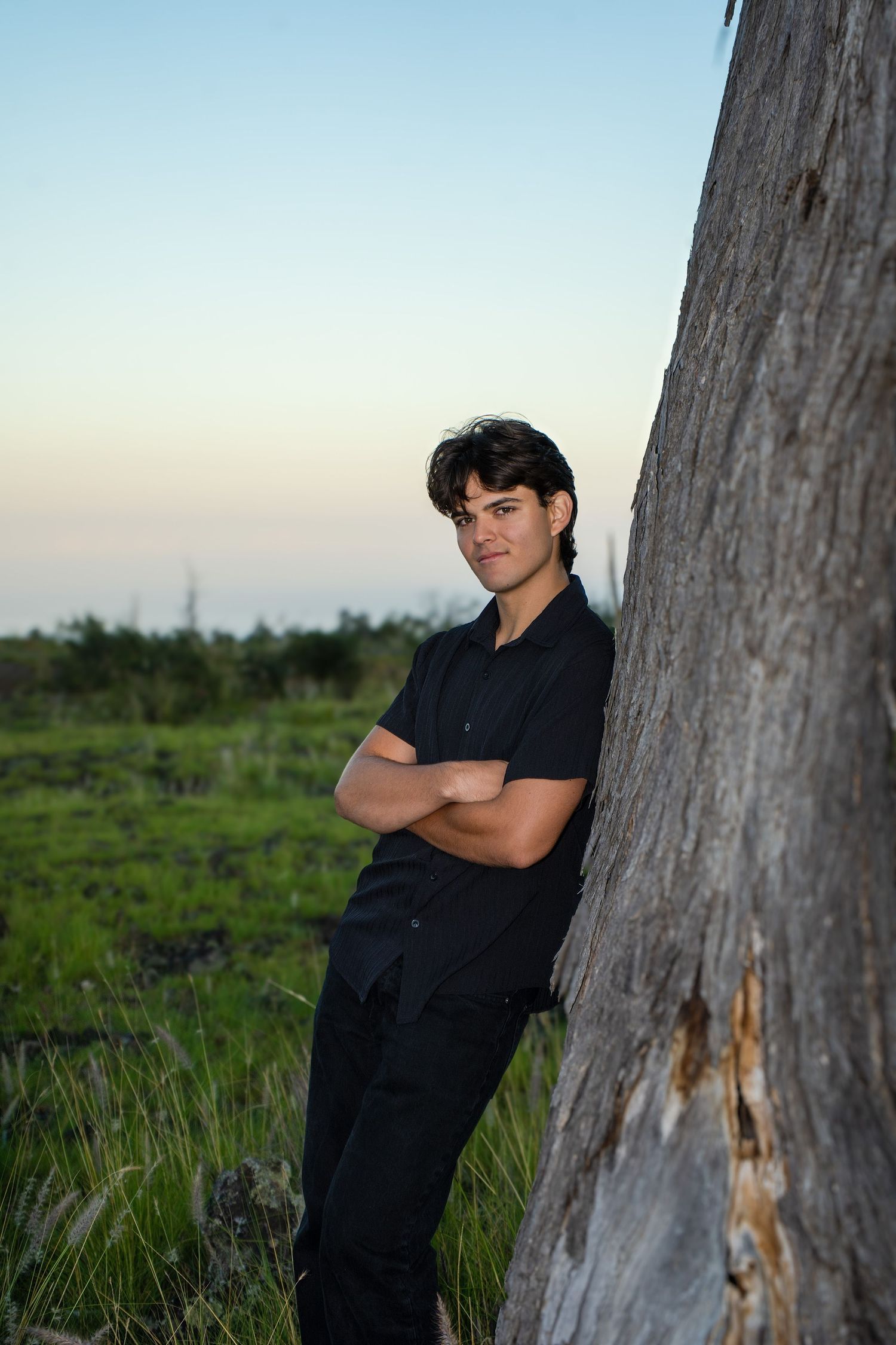 A figure in black clothing leans against a tree trunk in a grassy field at sunset.