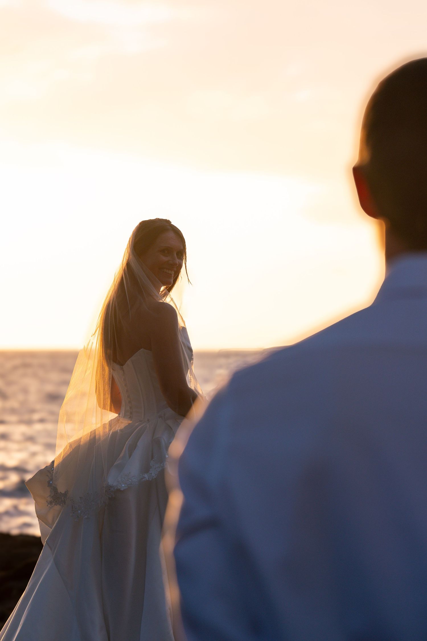 Bride in white gown and veil smiles at groom during romantic beach wedding at golden sunset.