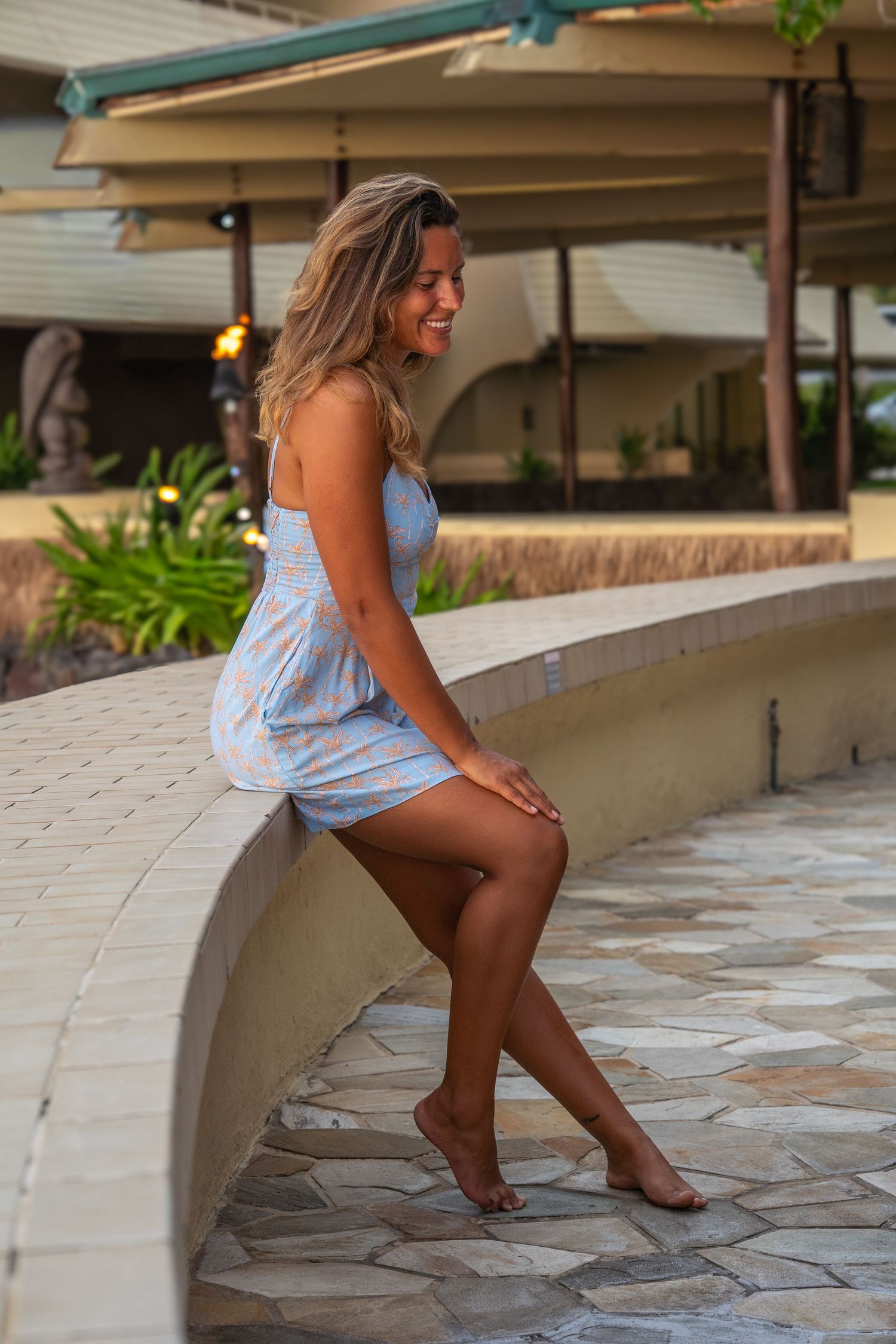 Smiling woman in light blue sundress sitting barefoot on stone wall at tropical resort outdoor area.