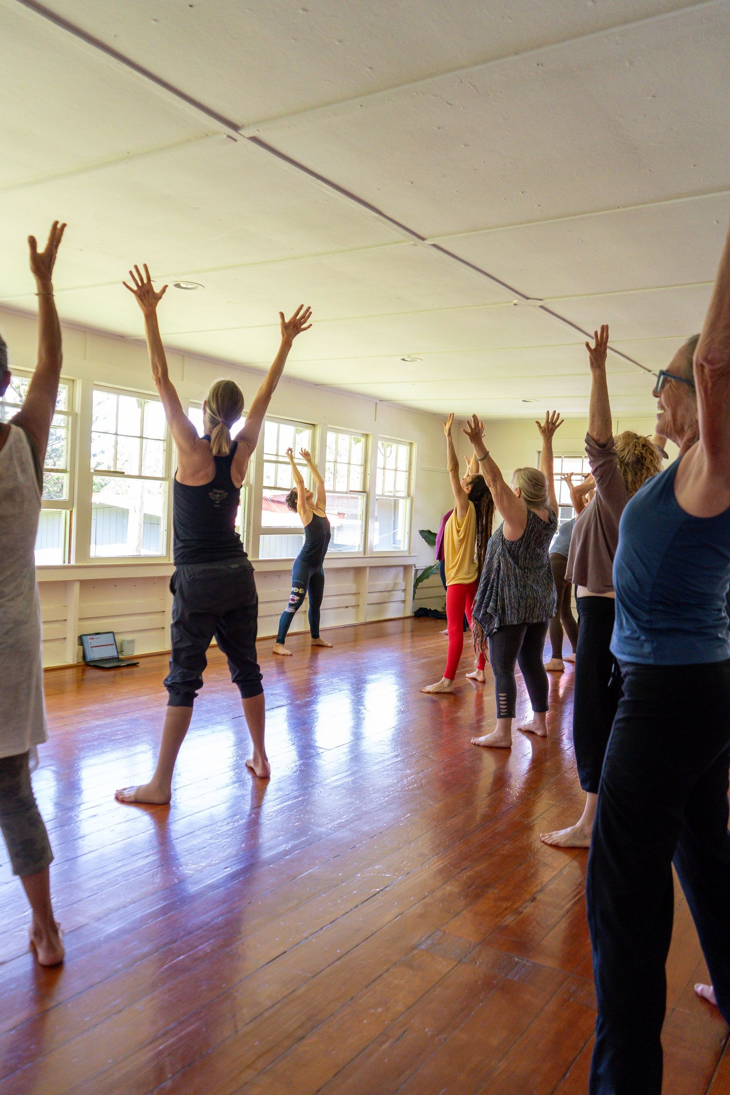 A group of people practicing yoga stretches with raised arms in a bright studio with wooden floors.