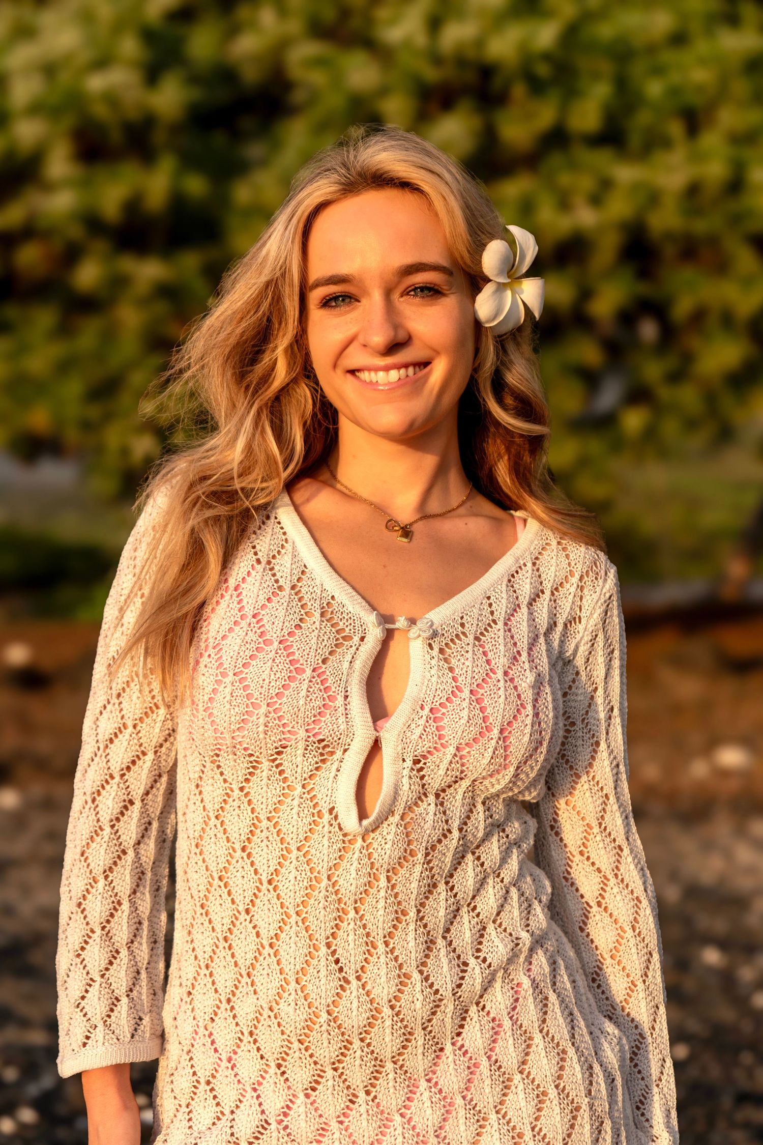 A person wearing a white crochet sweater smiles outdoors in natural lighting with a flower in their hair.