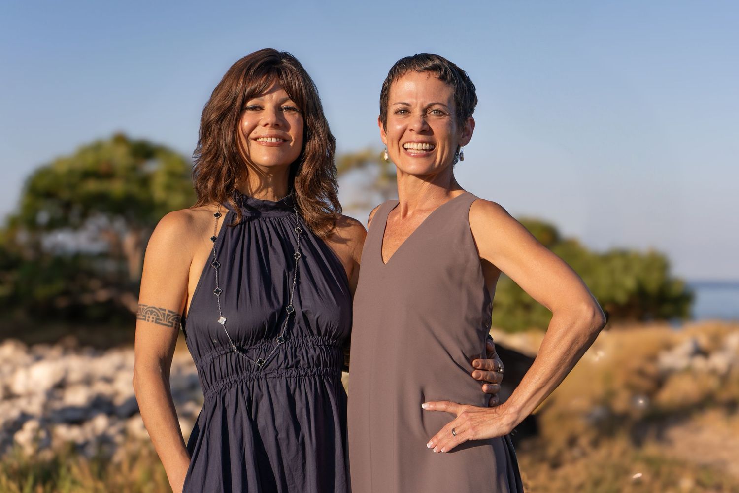 Two people in dark dresses stand together smiling on a coastal path during golden hour.