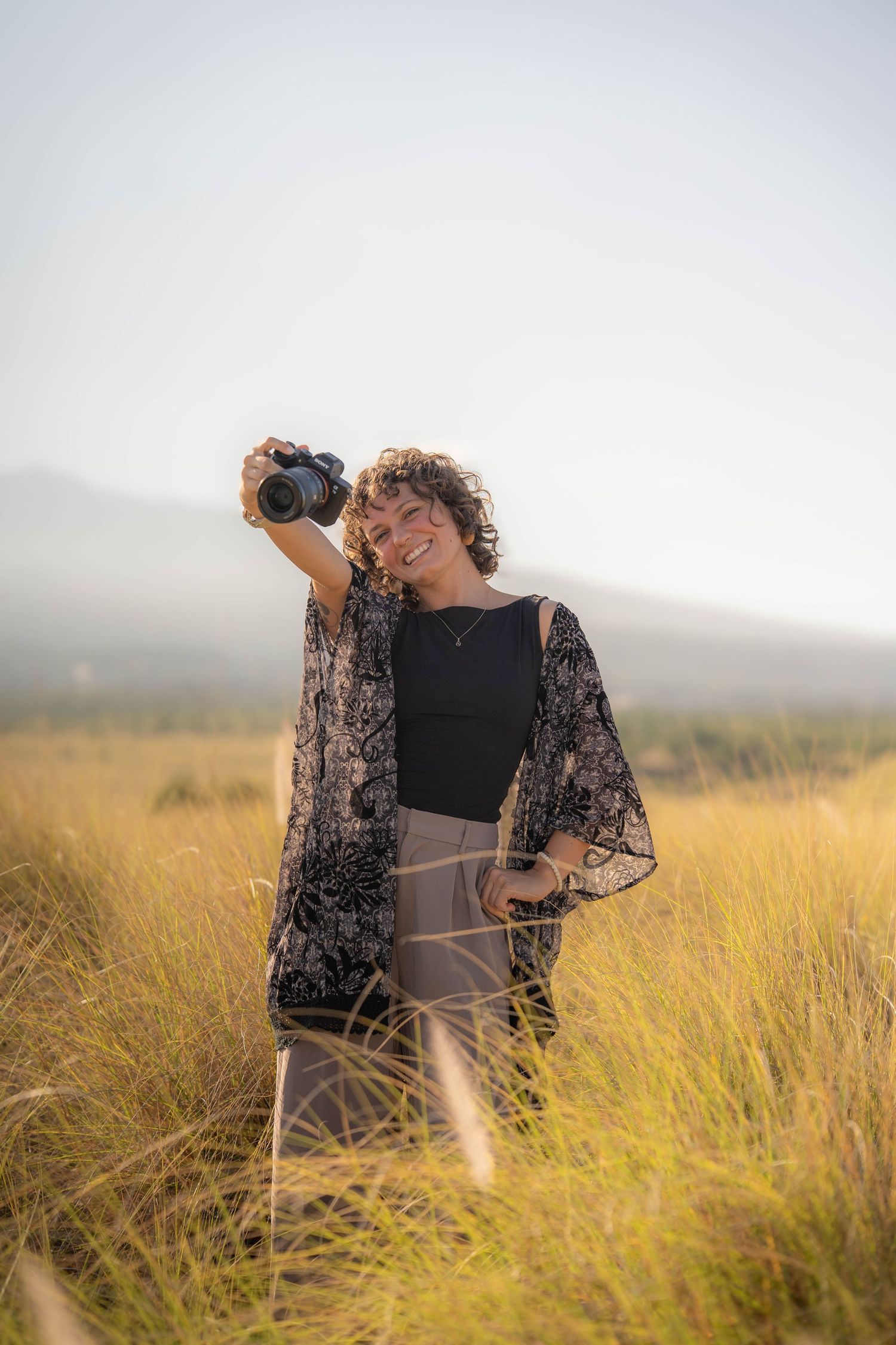 Smiling woman with curly hair holding a camera up in a golden grass field with mountains in the background.