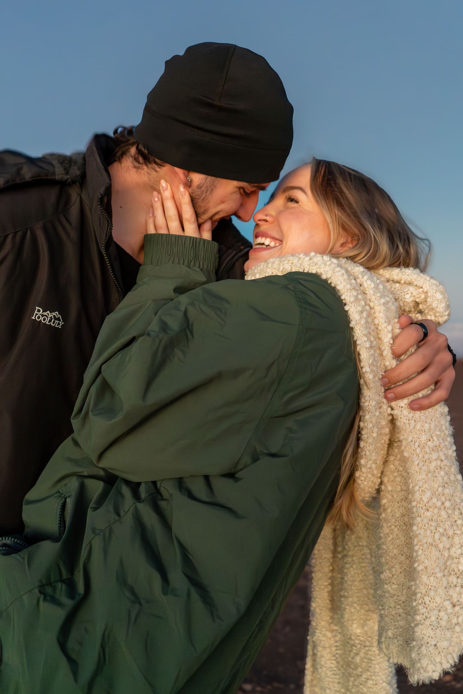 A couple in winter jackets share a romantic moment outdoors at sunset, smiling and embracing closely.