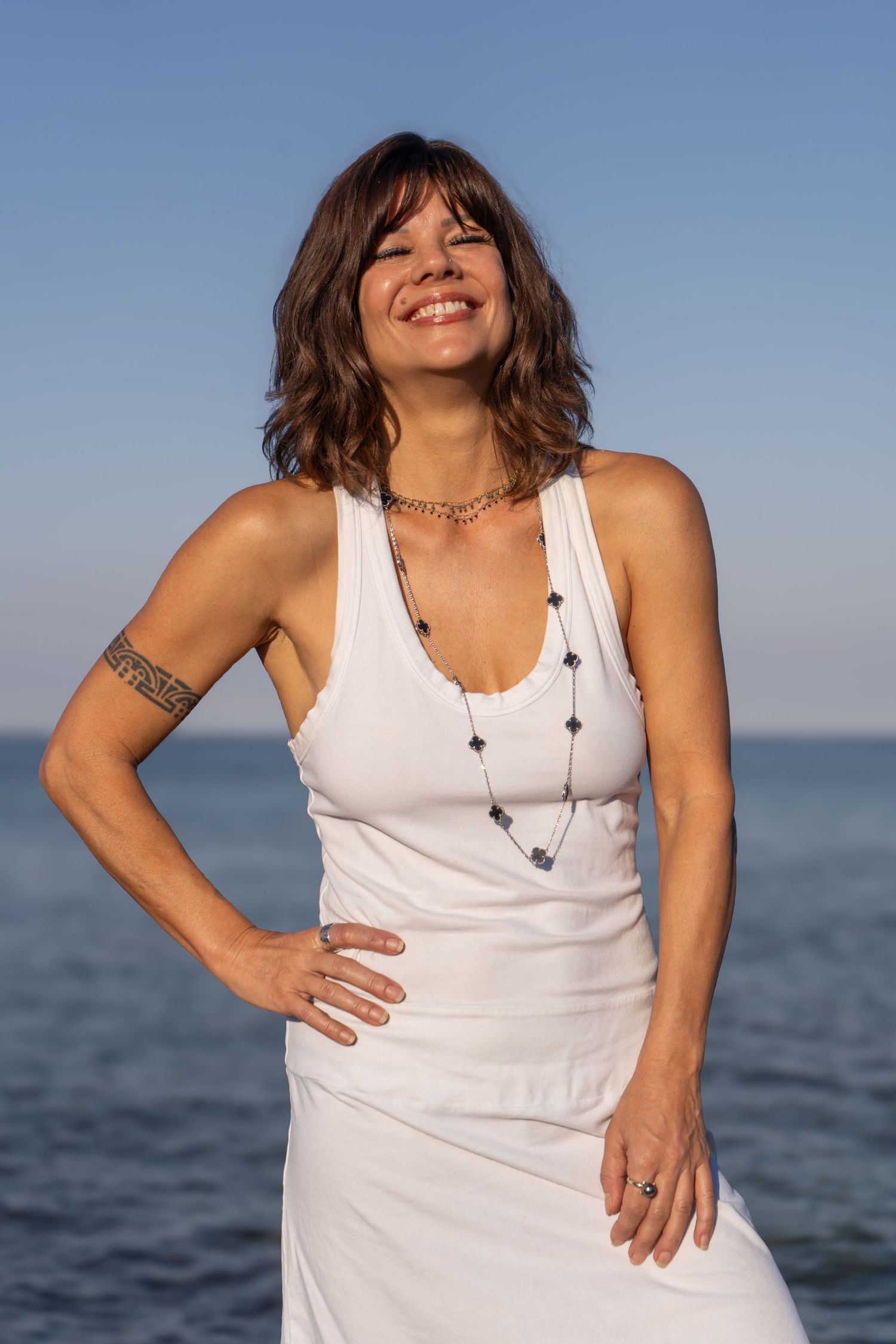 Person in a white tank top poses confidently by the ocean under blue skies.