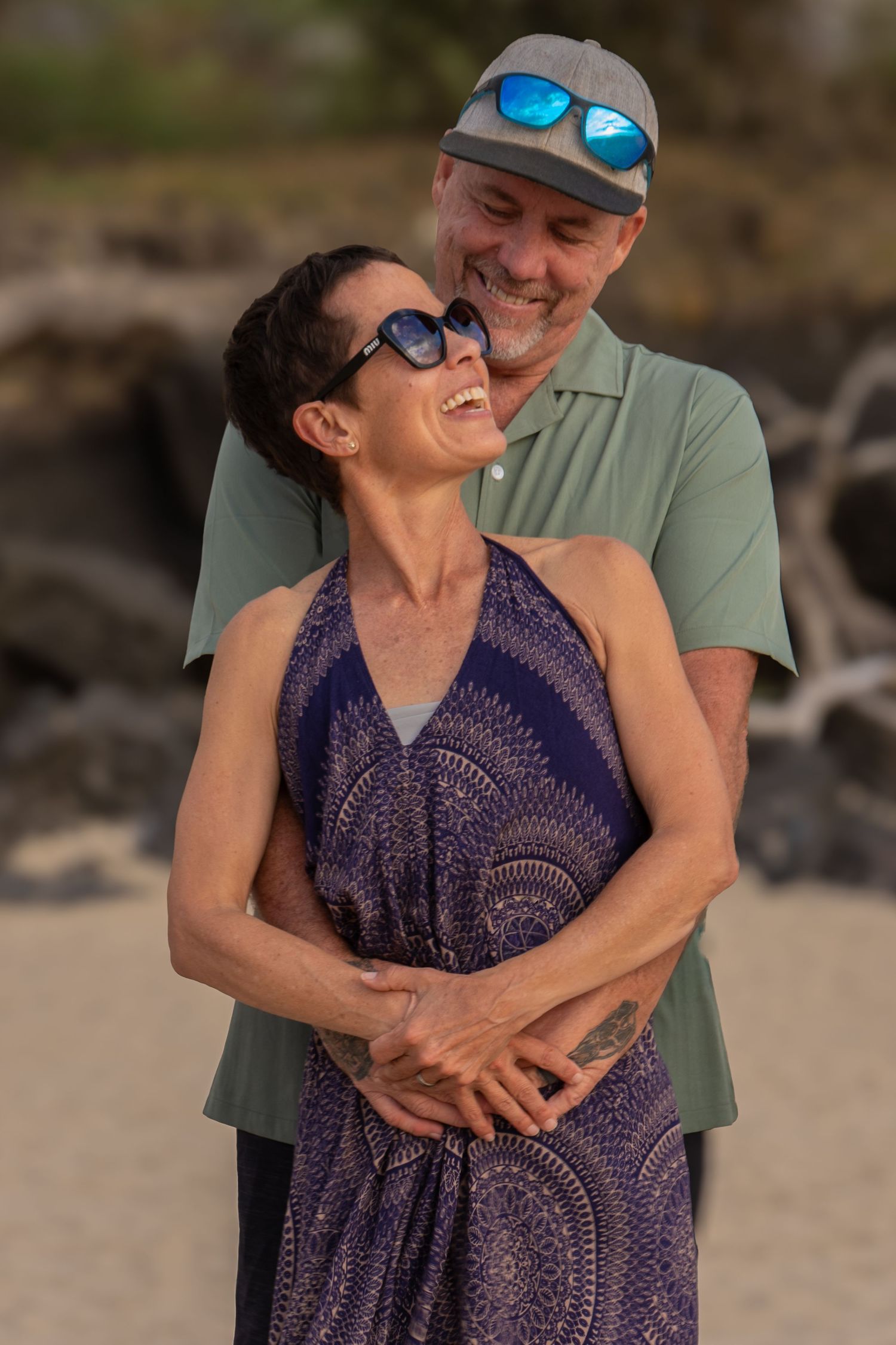 Couple embracing on beach at sunset wearing casual summer attire.