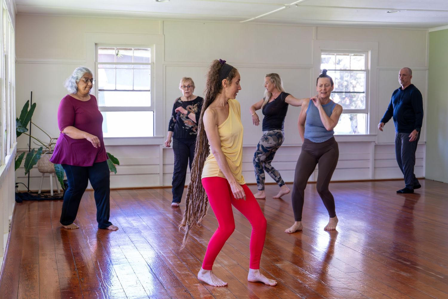 A group of people participate in a fitness class doing stretching exercises in a bright studio with wooden floors.