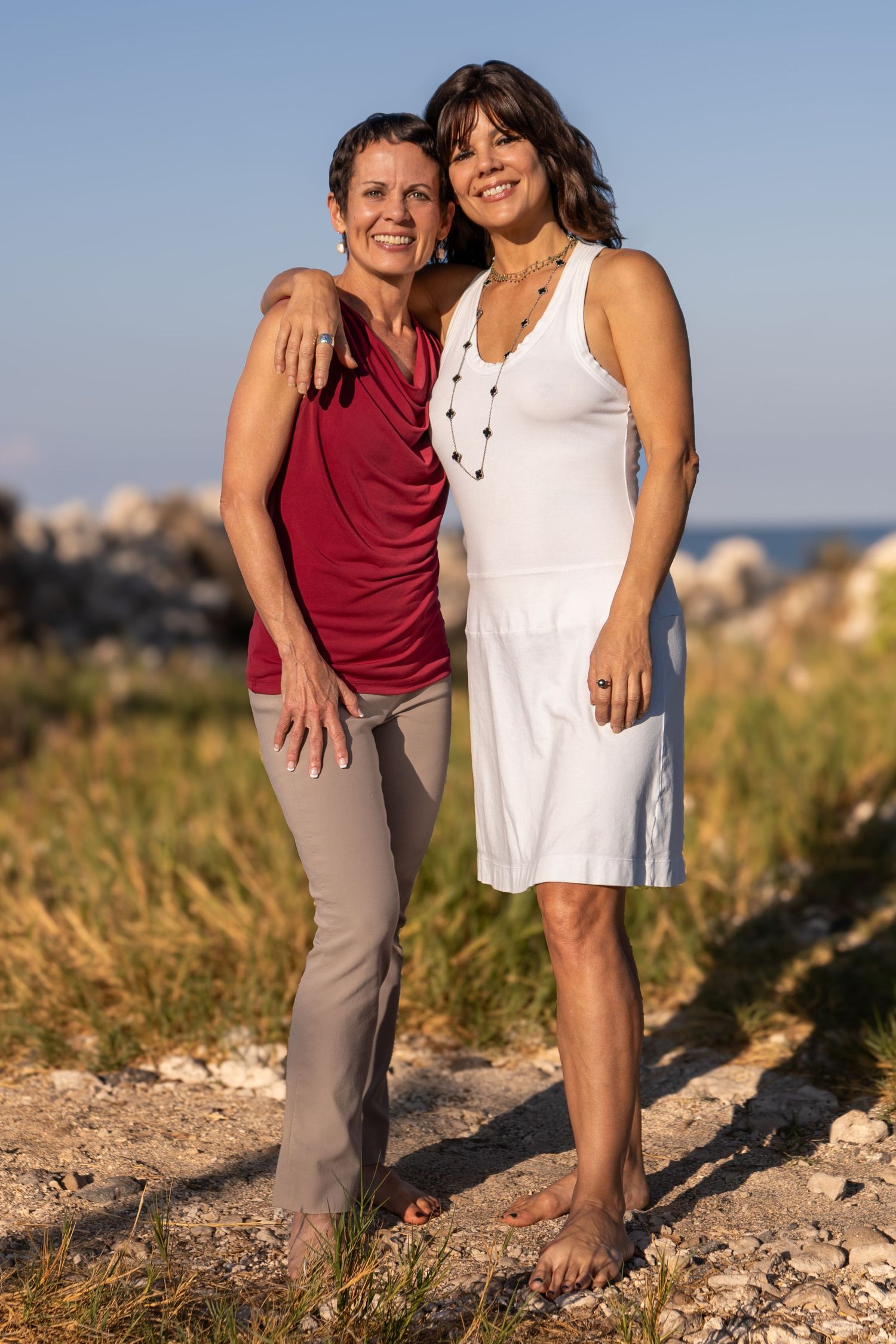 A couple stands together outdoors on a grassy hill during golden hour, wearing casual summer clothing.
