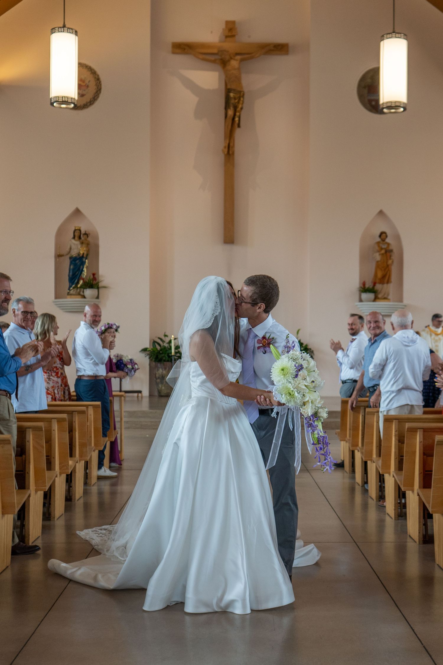 Bride and groom share first kiss at Catholic church wedding ceremony, guests standing in pews, large crucifix on wall.