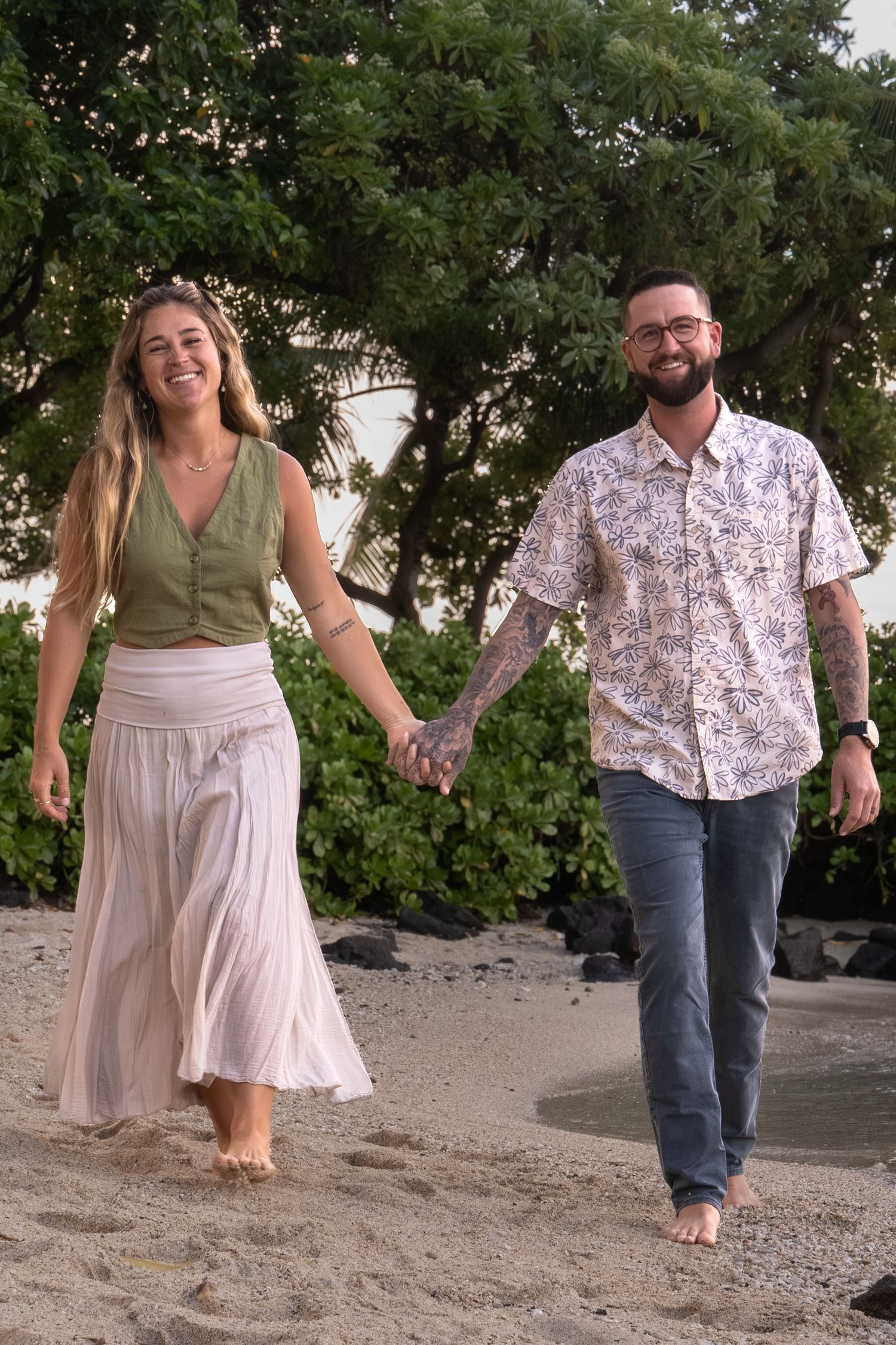 A couple dances and laughs together on a sandy beach with green trees in the background.