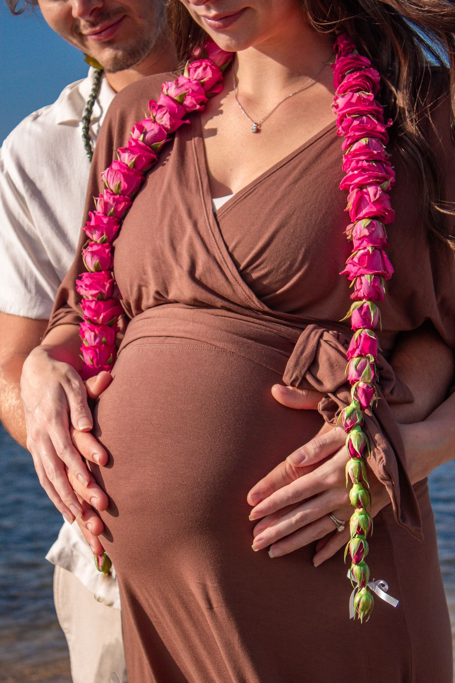 A person in a brown top wears a bright pink floral lei while standing near the ocean at sunset.
