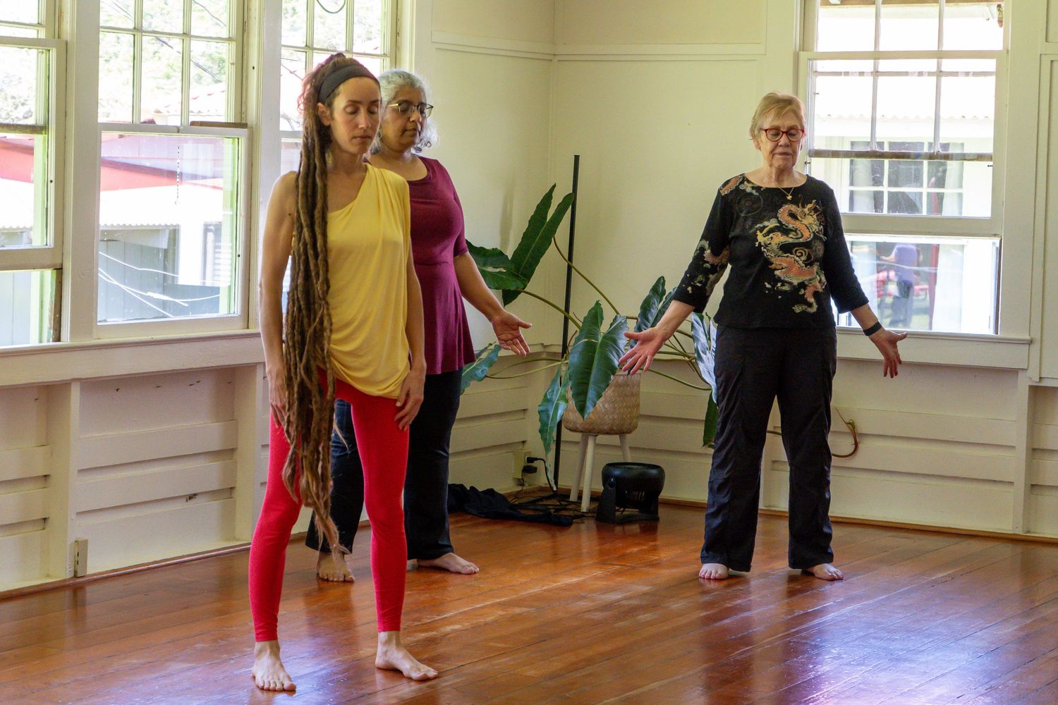 Three people practice dance moves together in a bright sunlit studio room with wooden floors and large windows.