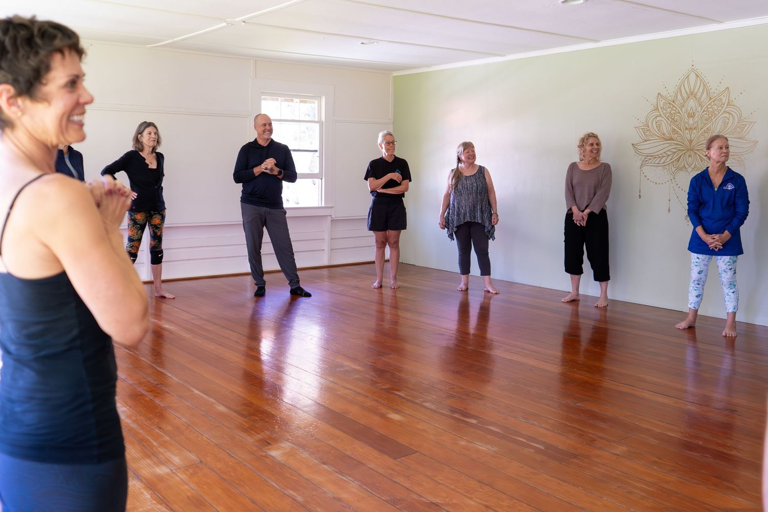 A group of people stand in formation during a dance or exercise class in a bright studio with wooden floors.