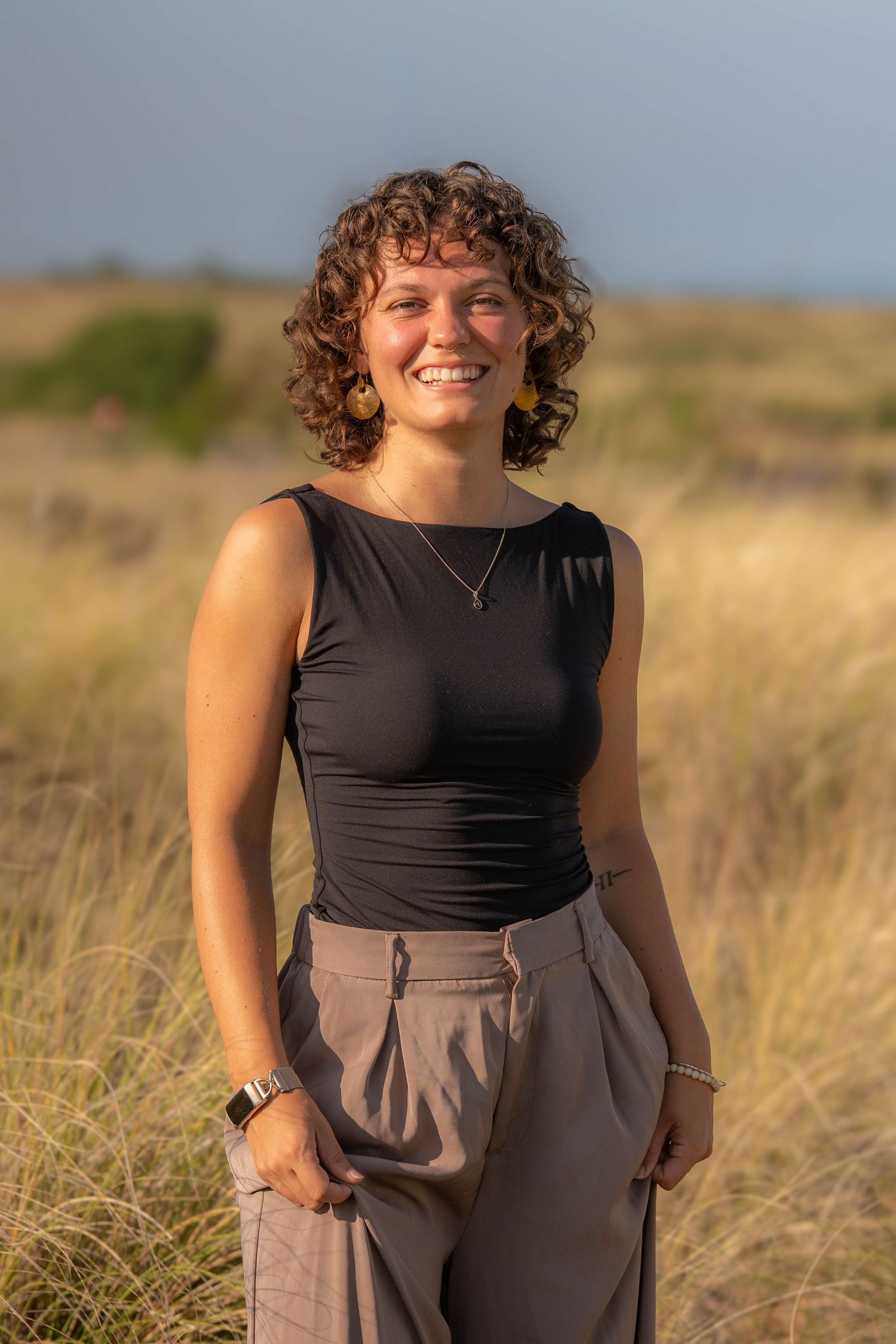 Smiling woman with curly hair in black sleeveless top and tan trousers standing in golden grass field at sunset.