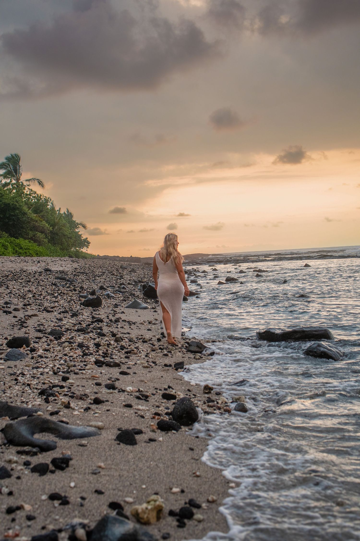 Woman in white dress walking on rocky beach at sunset with palm trees and dramatic cloudy sky.